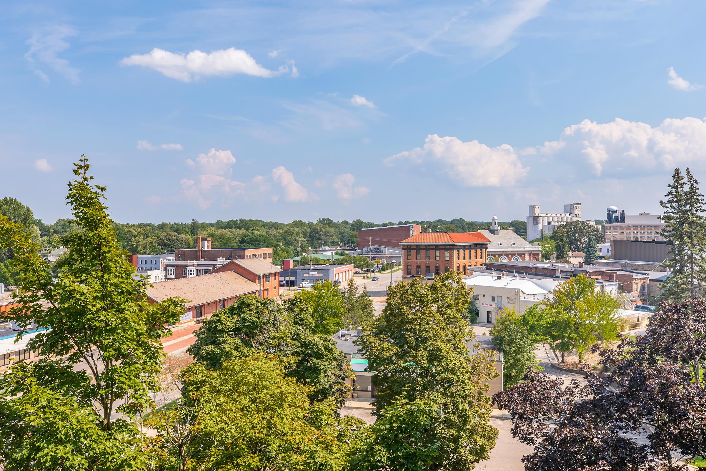 A scenic view of a small town with a mix of historic and modern buildings, framed by green trees. The sky is partly cloudy, and the landscape showcases a blend of urban and natural elements, with a hint of bustling activity in the streets below.
