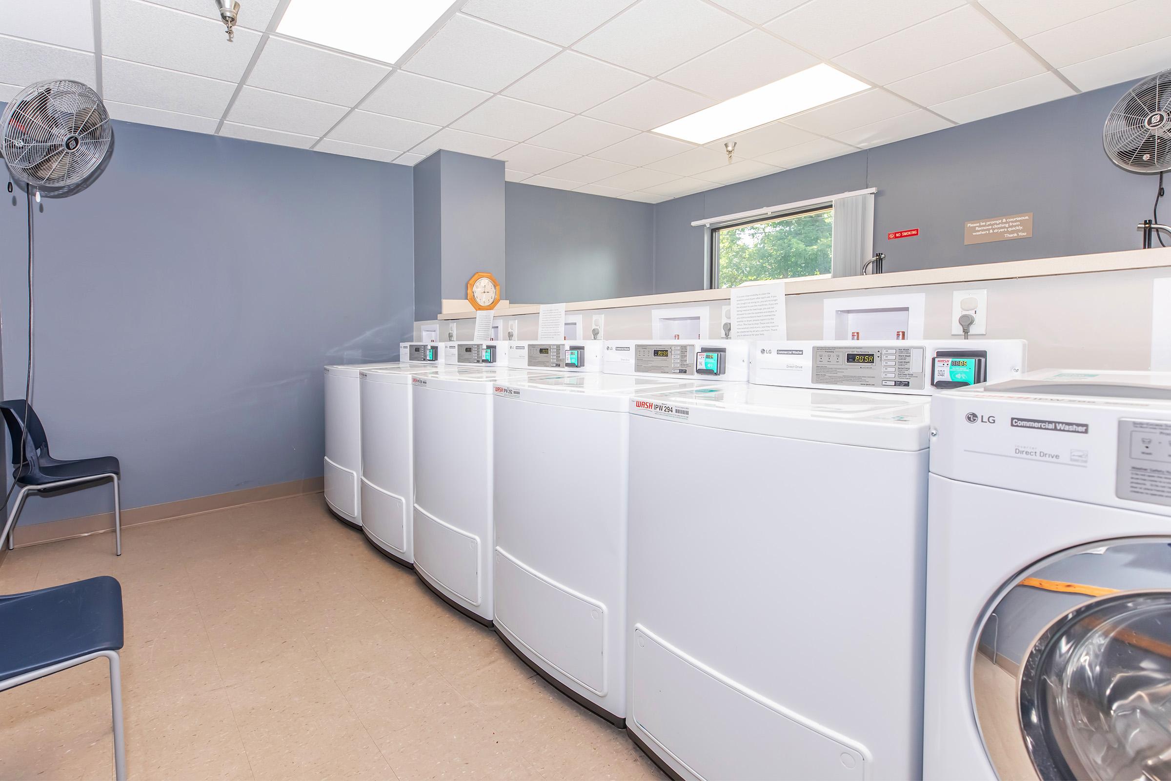A laundry room featuring several white washing machines lined up against the wall, with a large window providing natural light. The floor is tiled, and there are two fans on the ceiling. A simple wall clock and a window curtain can be seen, creating a clean and organized space for doing laundry.