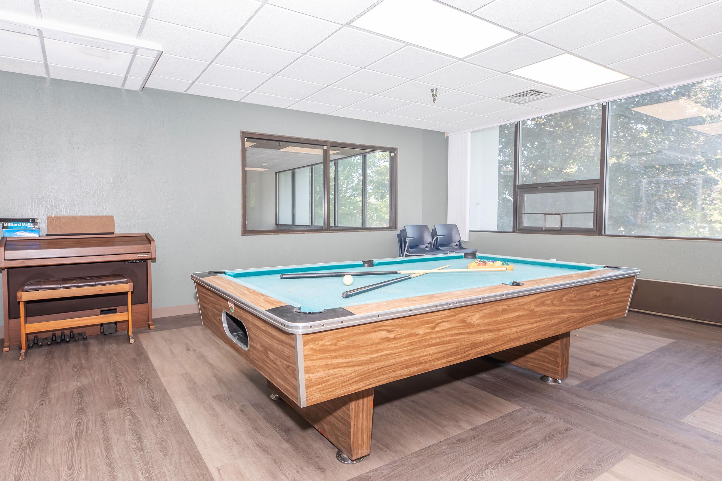 A well-lit game room featuring a pool table with cues and balls. There are two blue chairs in the background, a piano in the corner, and large windows allowing natural light. The room has wood and laminate flooring, creating a relaxing atmosphere for leisure activities.