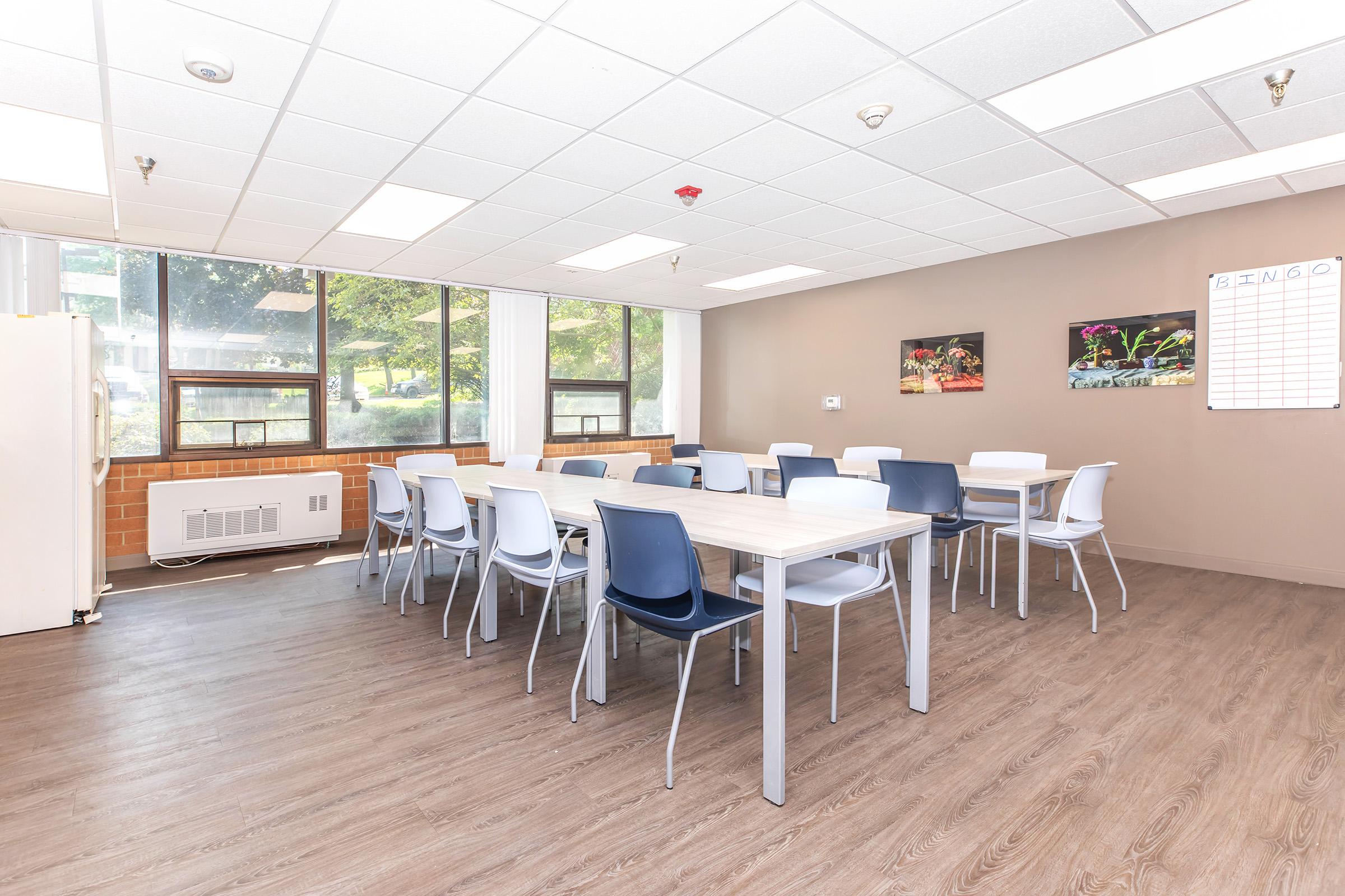 A spacious and well-lit classroom with large windows, featuring a long table surrounded by chairs. There are two framed pictures on the wall, a refrigerator in the corner, and a whiteboard with a BINGO grid. The floor is made of wood laminate, creating a warm and inviting atmosphere.