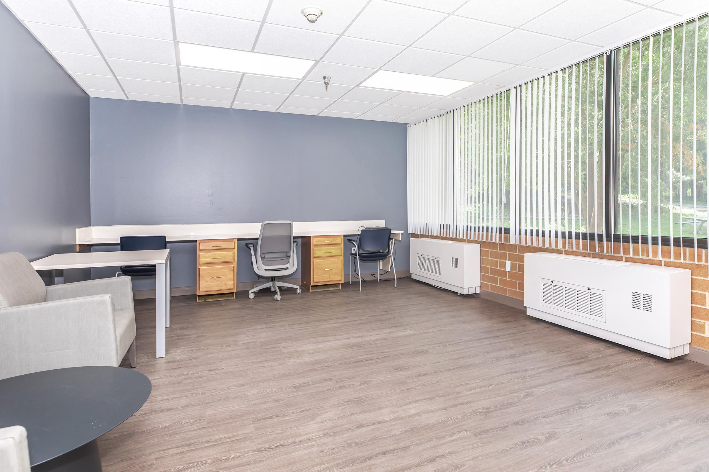 A modern, spacious office space featuring a gray sofa, a round black table, and two desks with chairs. Large windows with vertical blinds let in natural light, illuminating the hardwood floor and painted blue walls. The minimalist design creates an open and inviting atmosphere.