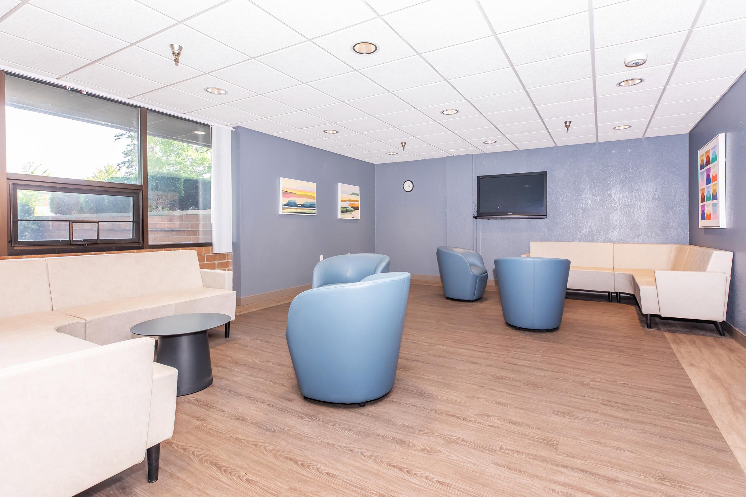 A modern lounge area featuring light-colored sofas and blue armchairs arranged around a circular black coffee table. The walls are painted in a soft blue with framed art prints. A wall-mounted television and a clock can be seen, and large windows offer natural light. The floor is wooden, creating a warm atmosphere.