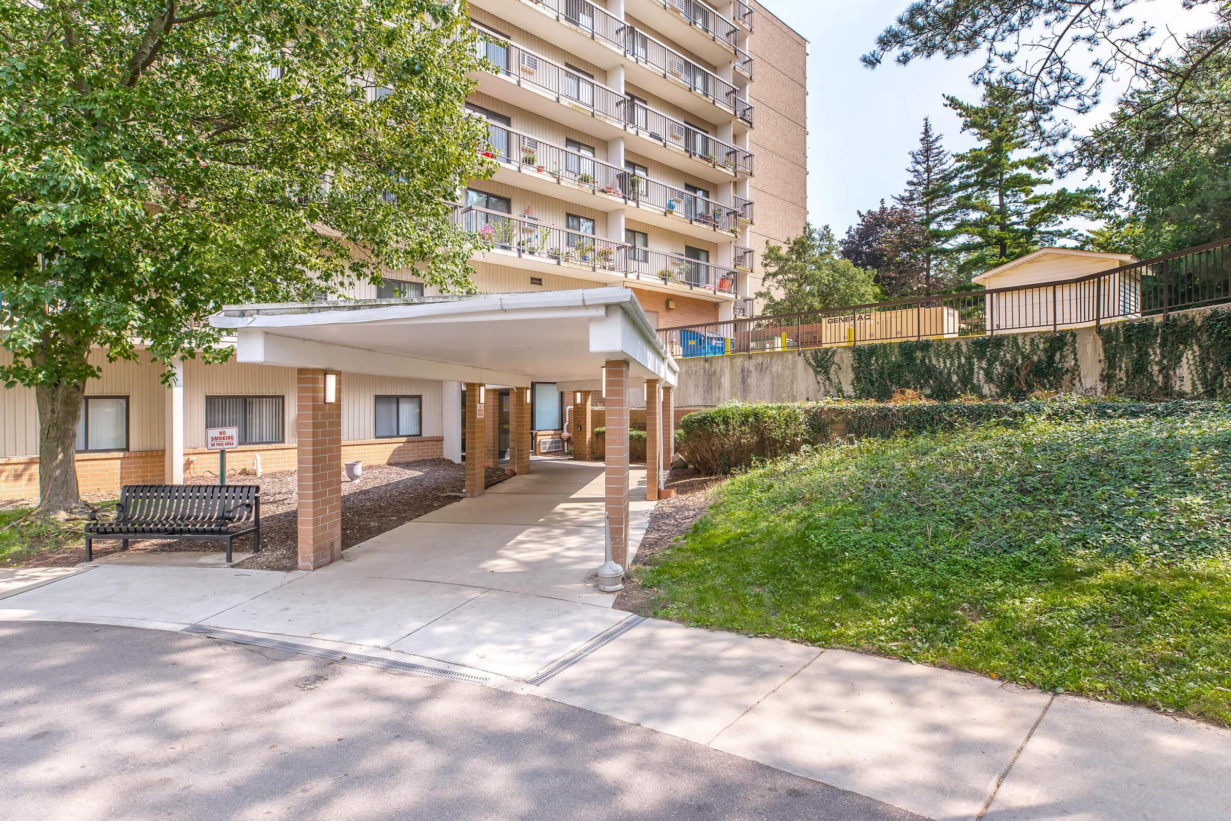 A view of an apartment building entrance featuring a covered walkway and a bench. Lush greenery surrounds the area, with a well-maintained lawn and trees nearby. The building has multiple stories and balconies, reflecting a residential environment.