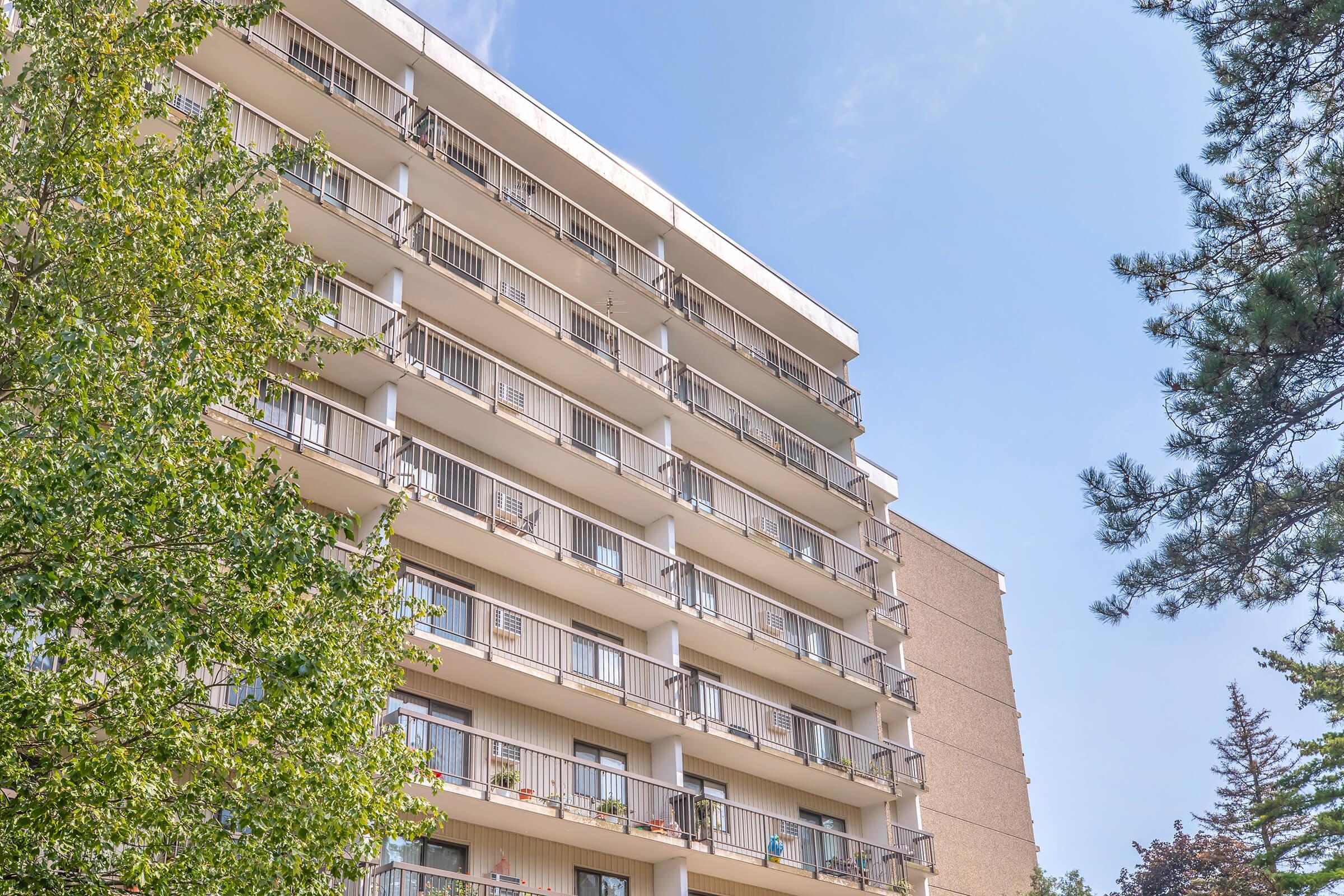 A multi-story residential building with balconies, featuring large windows and air conditioning units. Surrounding greenery includes trees, providing a natural contrast to the structure under a clear blue sky.