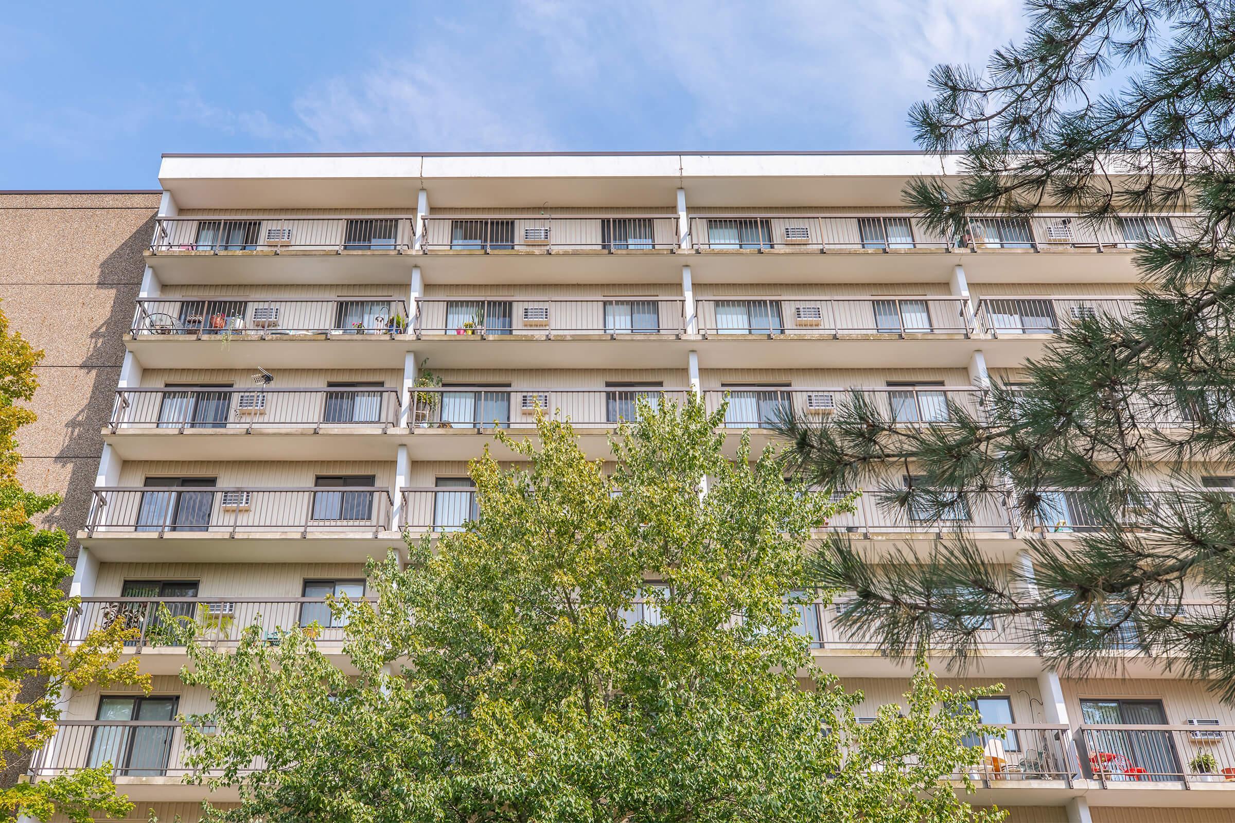 A modern apartment building with multiple balconies. The structure has four stories and is partially surrounded by trees. Several balconies display potted plants and personal items, while blue skies and scattered clouds are visible above.