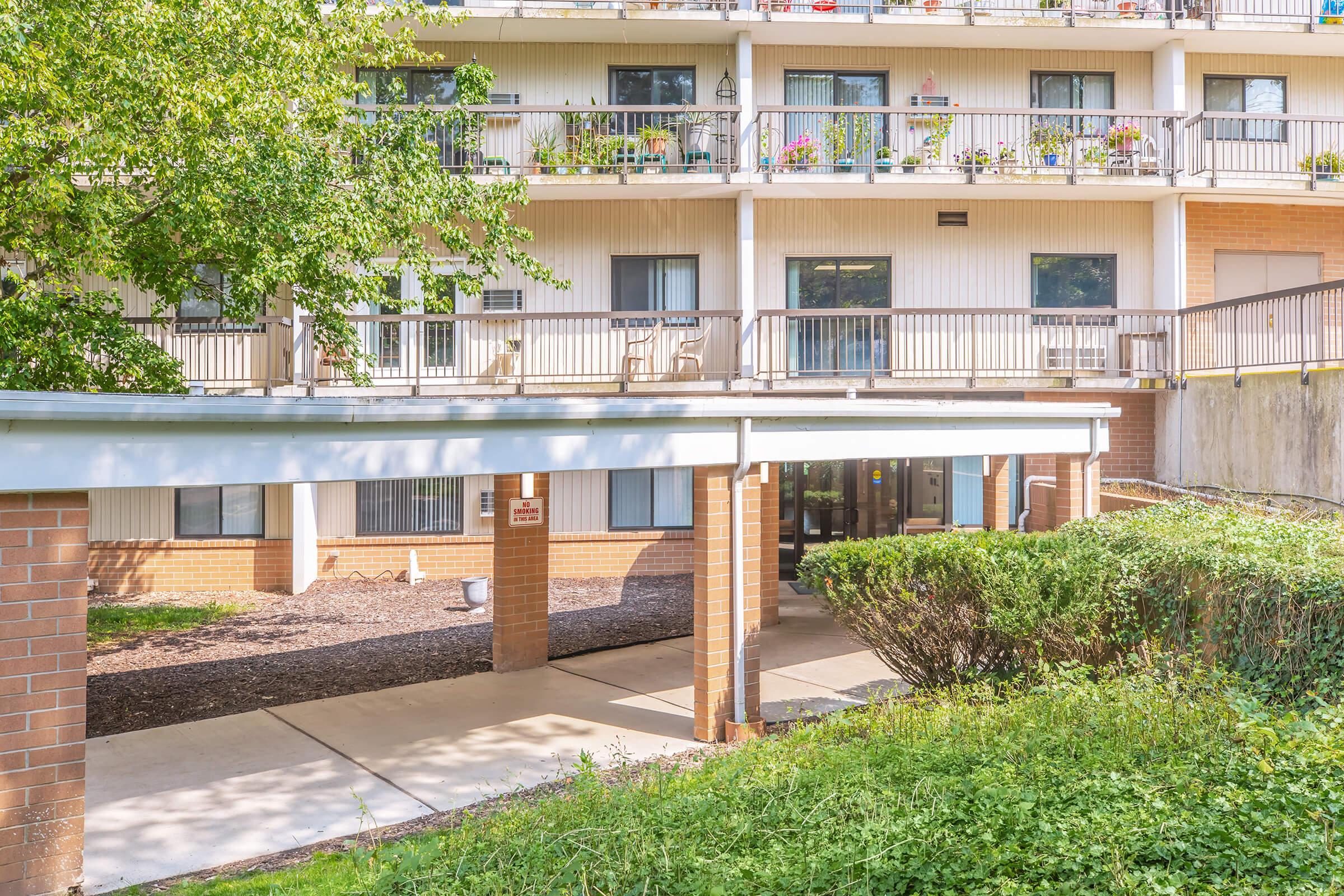 A view of a multi-story building with a covered entrance and outdoor seating area. The building features balconies adorned with greenery and flowers. There is a pathway leading to the entrance, surrounded by shrubs and grass. The scene is well-lit with natural sunlight.