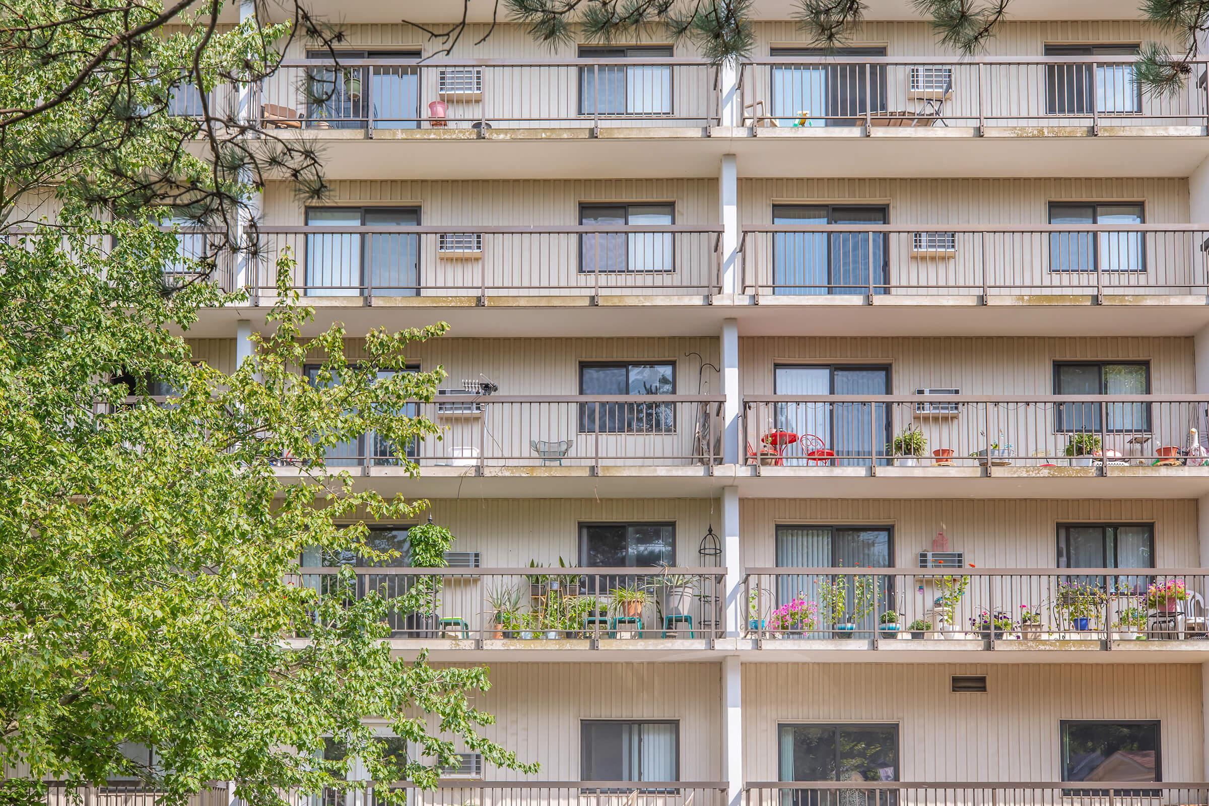 A multi-story residential building featuring several balconies decorated with potted plants and chairs. The structure has a light-colored exterior, with windows and air conditioning units visible. Green trees in the foreground provide a natural setting.