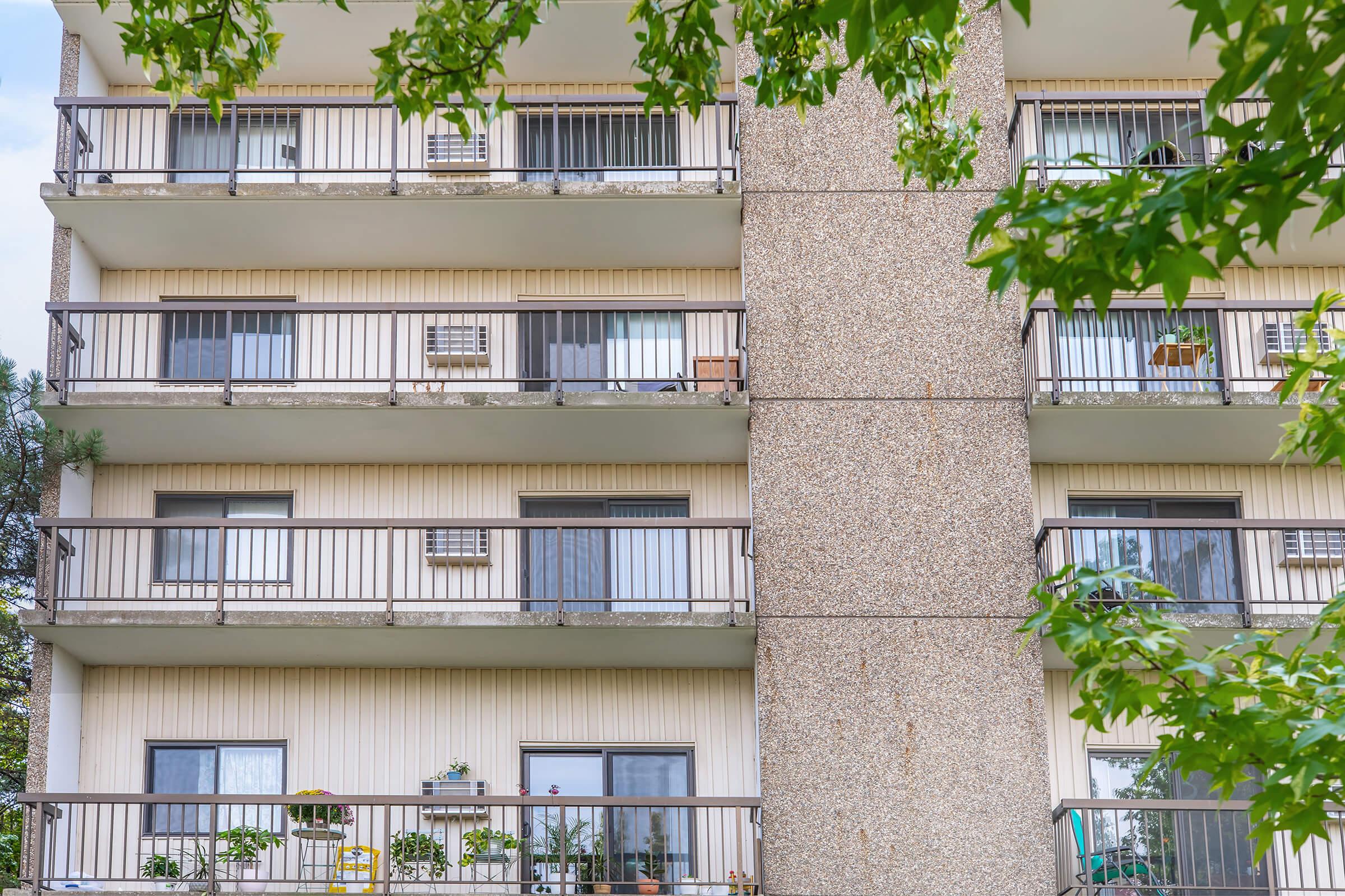 A close-up view of a multi-story residential building featuring several balconies with plants and seating. The building has a textured concrete column and light exterior walls. Green leaves from a nearby tree are partially visible in the foreground, adding a touch of nature to the urban scene.