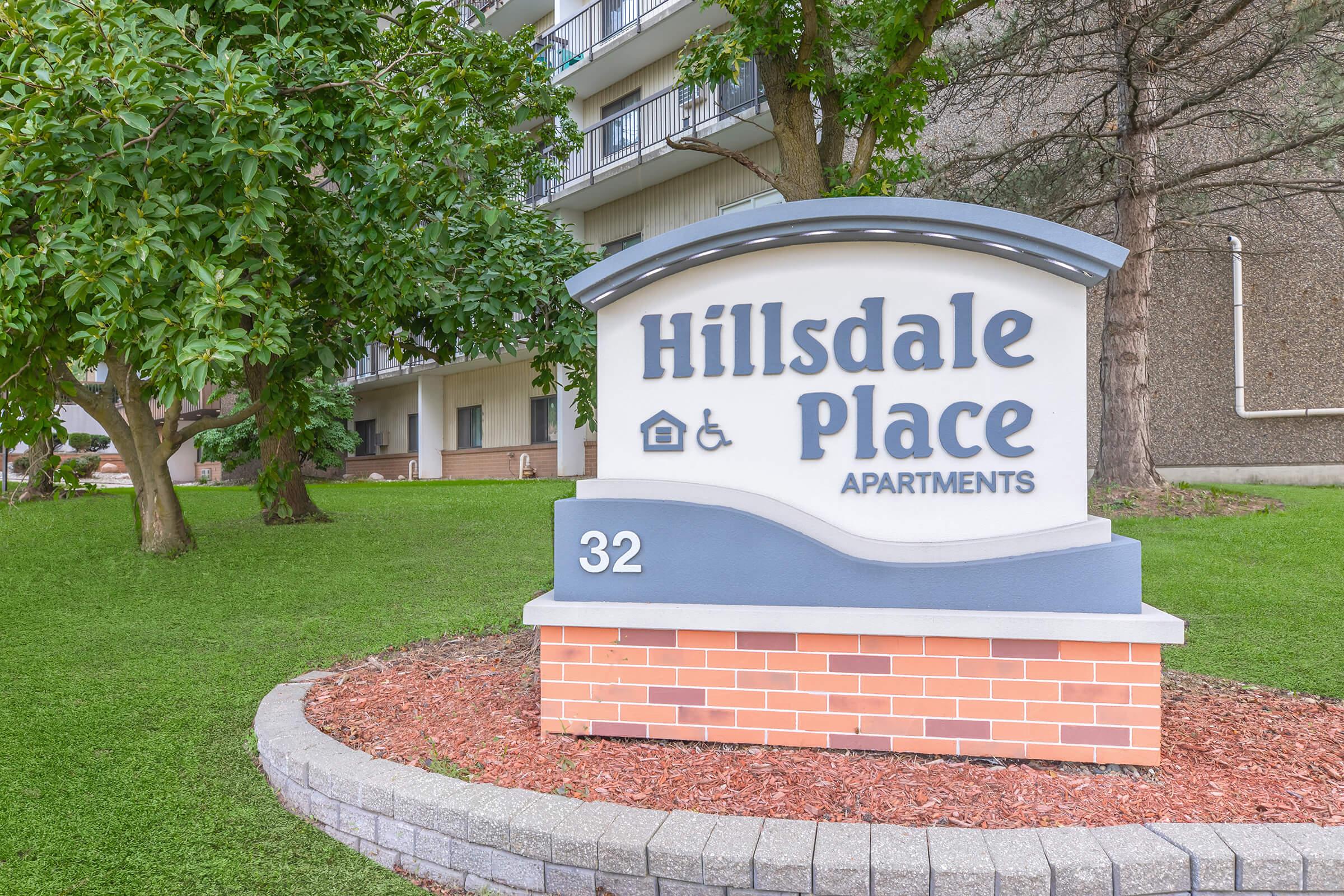 Sign for Hillsdale Place Apartments, featuring the name prominently at the top. The sign includes an icon for accessibility and the number 32. It is situated in a landscaped area with grass and trees in the background, showcasing a welcoming entrance to the apartment complex.
