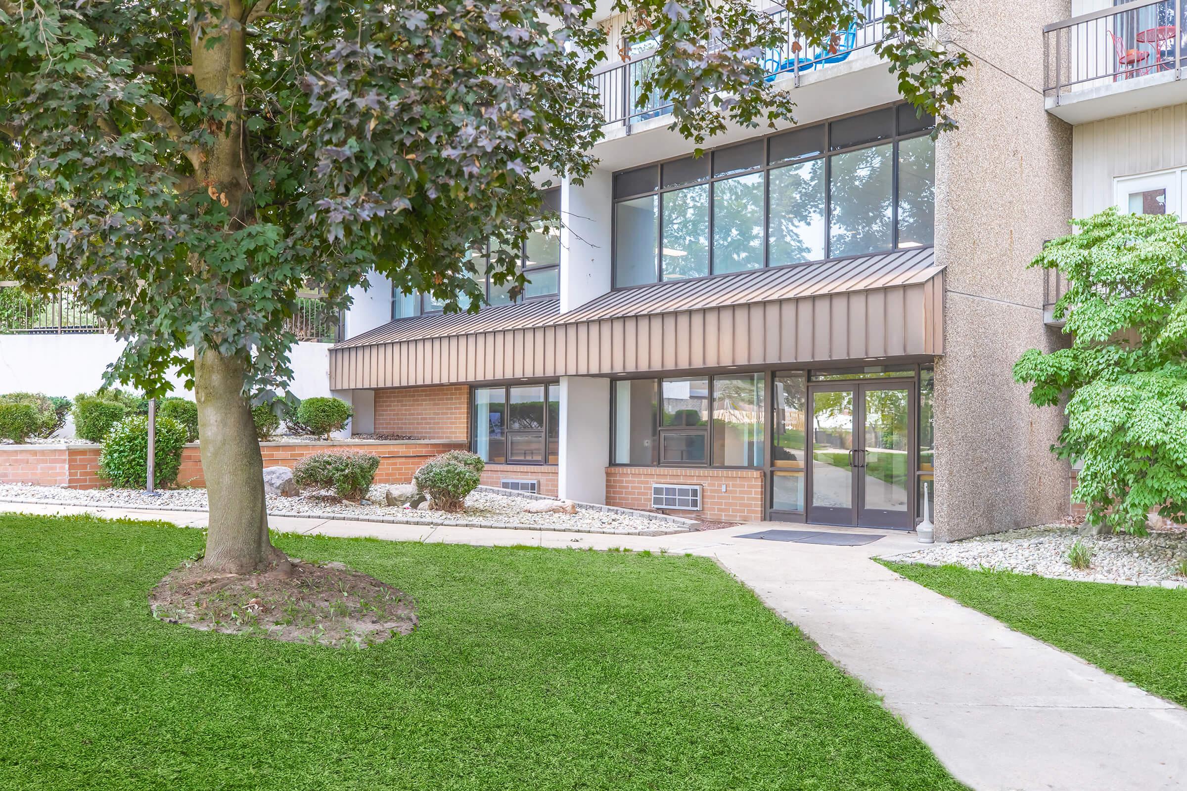 Exterior view of a modern building featuring a glass entrance with a brown awning. The surroundings include well-maintained green grass, shrubs, and a tree. The pathway leads to the entrance, showcasing a clean and inviting atmosphere.