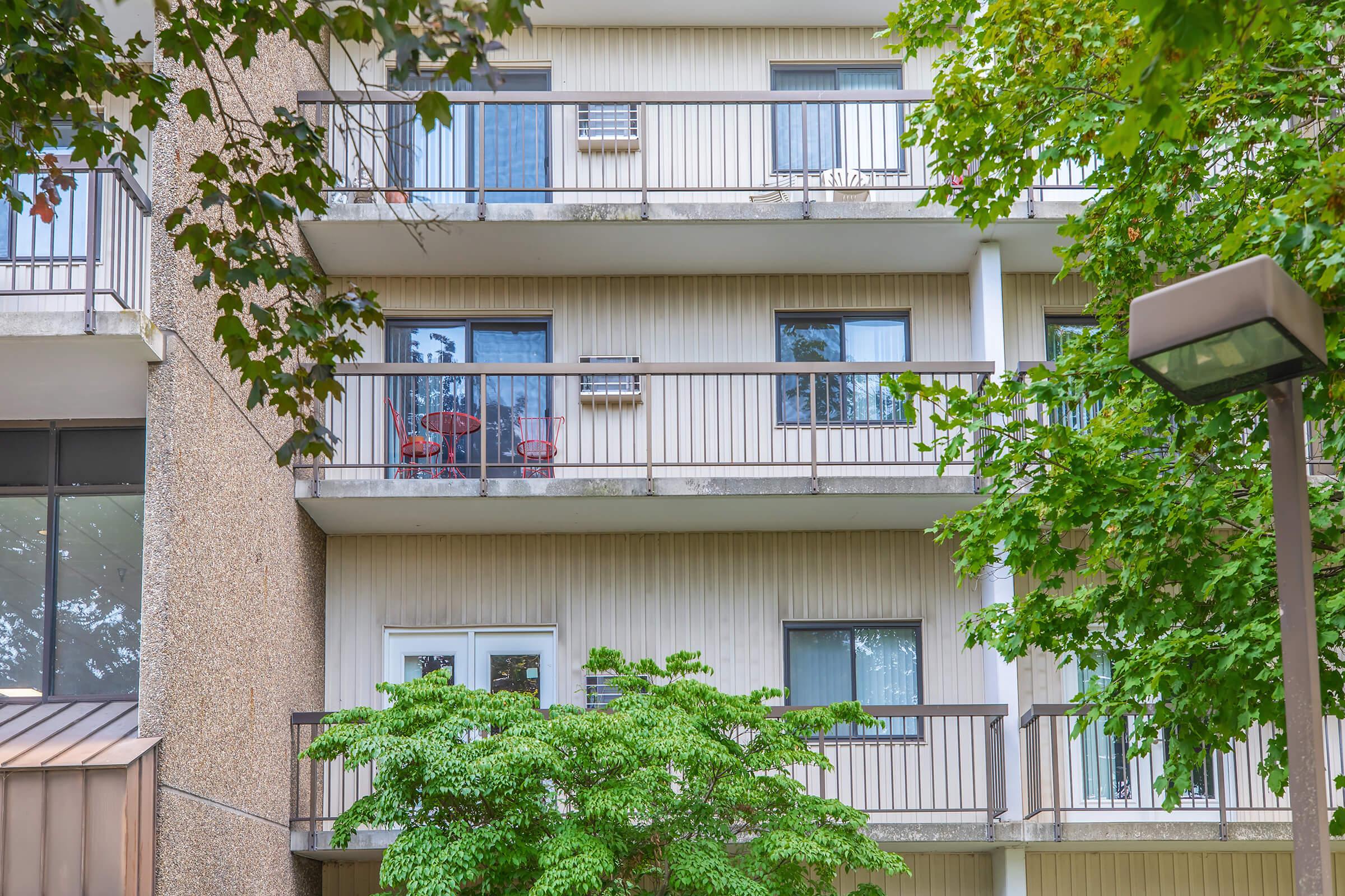 A multi-story building with balconies on each floor. Some balconies feature chairs and small tables, while trees and greenery surround the base of the building. The exterior has a textured surface, and windows are visible, some with air conditioning units.