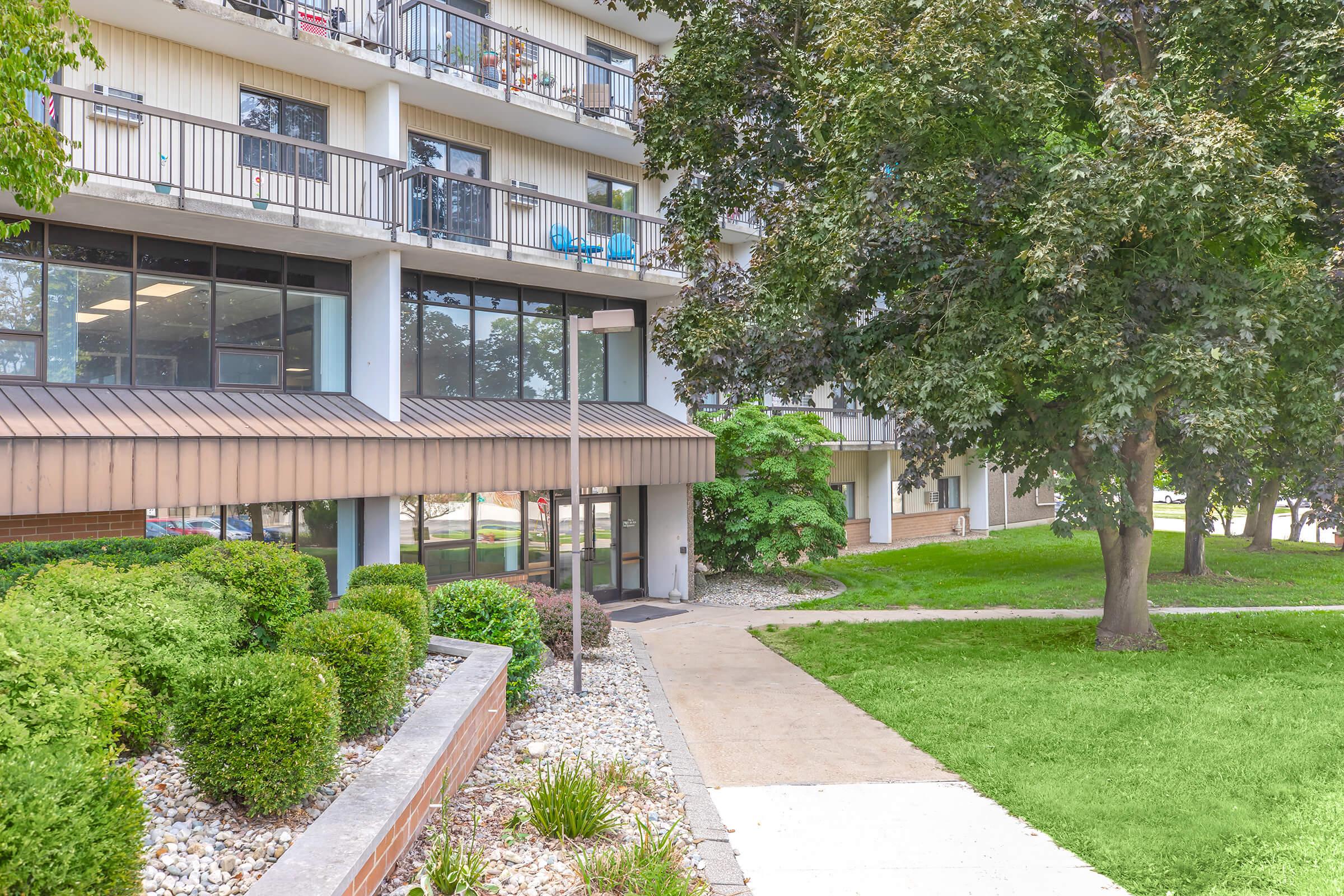 A view of a modern building with multiple stories, featuring a glass entrance and balconies. The surrounding area includes manicured greenery, shrubs, and a pathway leading to the entrance. The setting appears well-maintained and inviting, with a lush lawn and decorative stones.