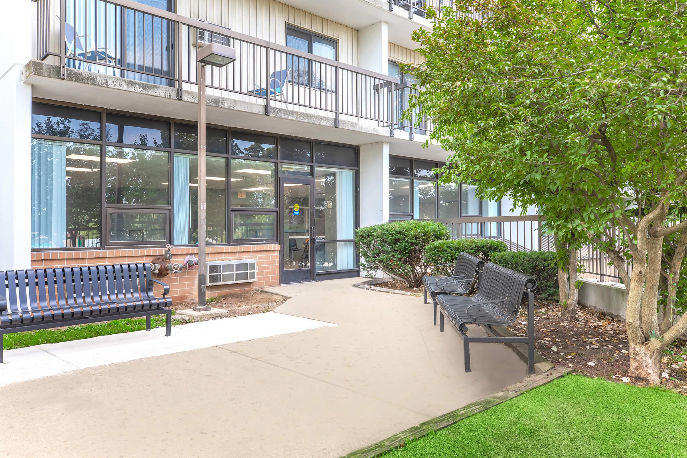 A view of a modern building exterior featuring benches, a well-maintained path, and lush greenery. The entrance has large windows allowing light in, with seating areas visible on the balcony above. The scene conveys a welcoming atmosphere for visitors and residents.