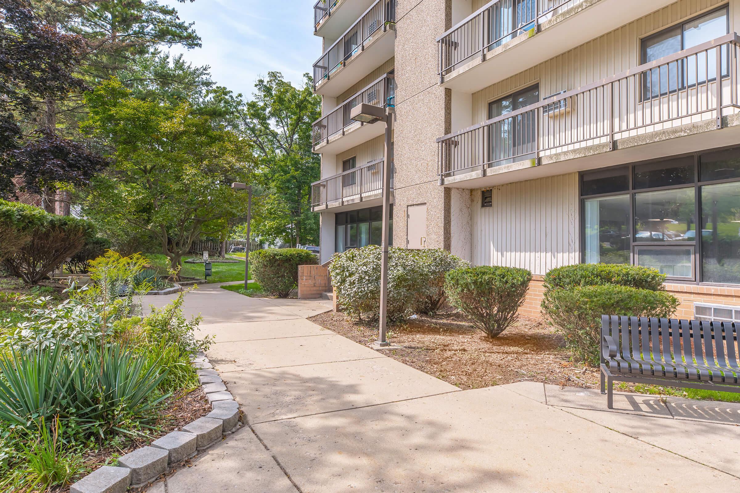 A well-maintained pathway leads through landscaped greenery towards a multi-story building. The area features bushes, trees, and a bench, with balconies visible on the building. Natural light brightens the scene, creating a welcoming environment.
