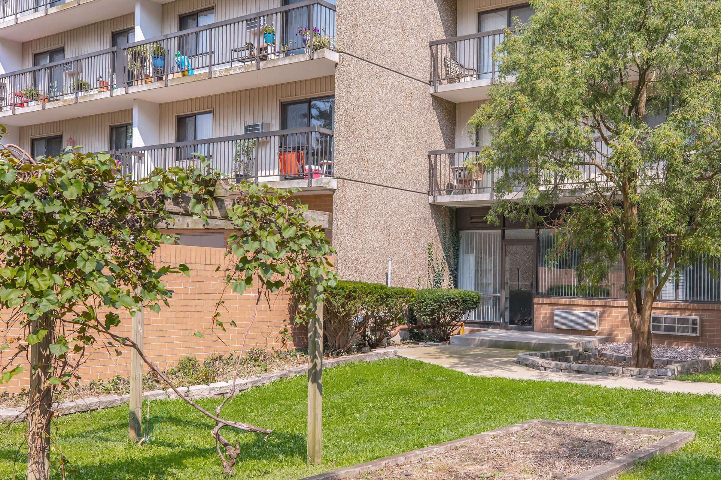 A multi-story apartment building with balconies adorned with potted plants. In the foreground, there is a garden area with green grass and several shrubs, along with a tree. The scene depicts a sunny day, showcasing the building's exterior and surrounding landscape.
