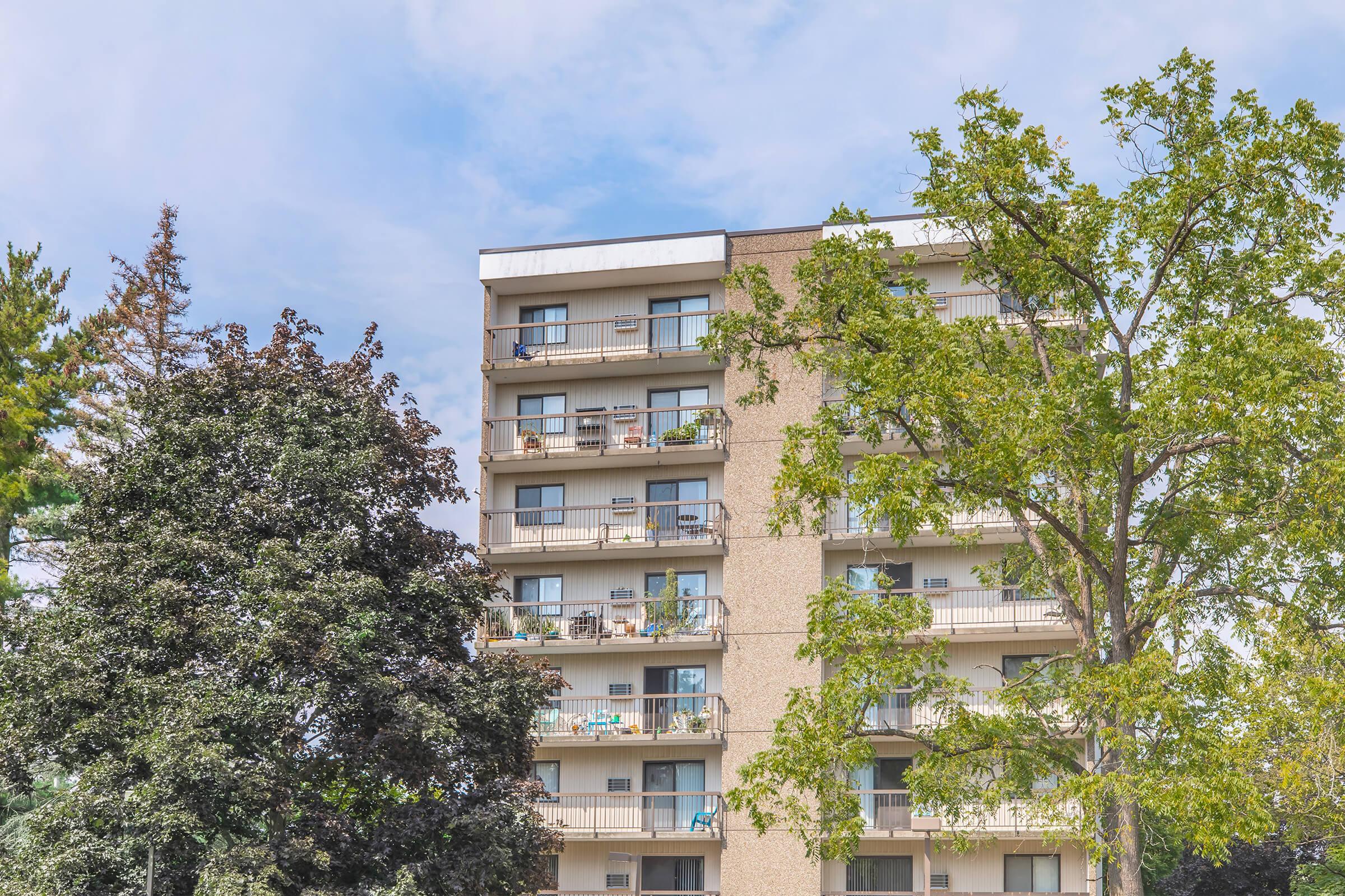 A multi-story apartment building with several balconies, some decorated with plants and outdoor furniture. Surrounding the building are various trees, including a large green one, and a clear blue sky with scattered clouds. The scene conveys a suburban residential atmosphere.