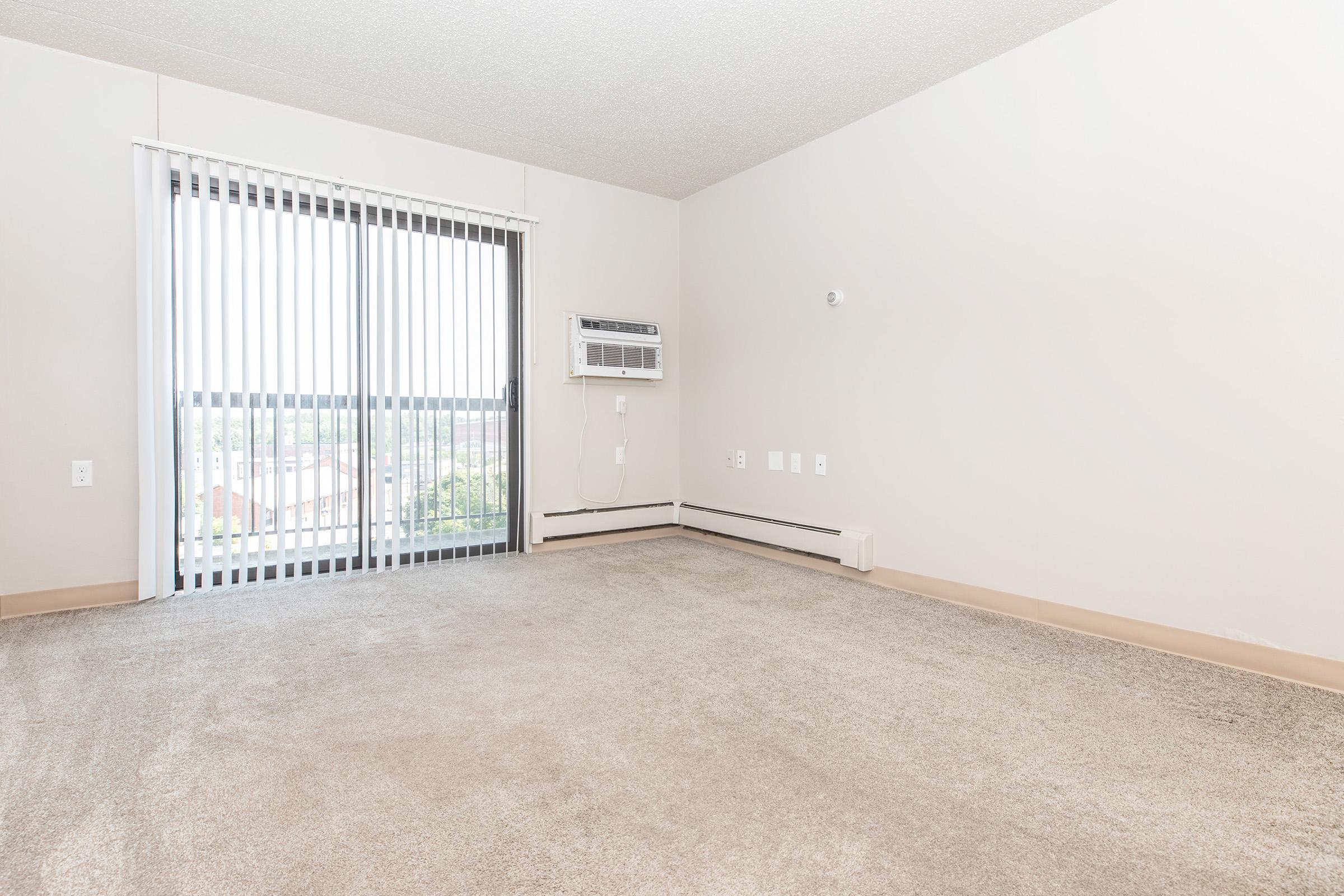 A spacious empty room with light-colored walls and a large window covered by vertical blinds. An air conditioning unit is mounted on the wall near the window, and the floor is carpeted in beige. There are visible electrical outlets and a neutral-colored baseboard along the wall.
