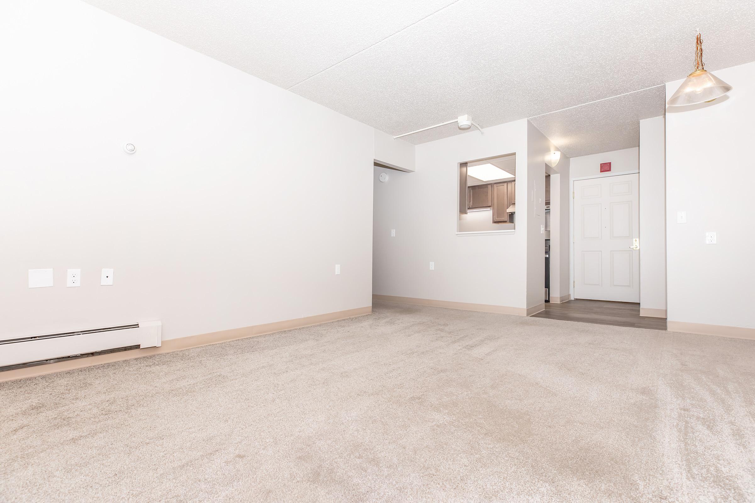 A spacious, empty living room featuring light gray carpet, neutral-colored walls, and a small kitchen area visible in the background. Soft lighting from a pendant light overhead enhances the open space, while a front door is situated to one side, creating a bright and inviting atmosphere.