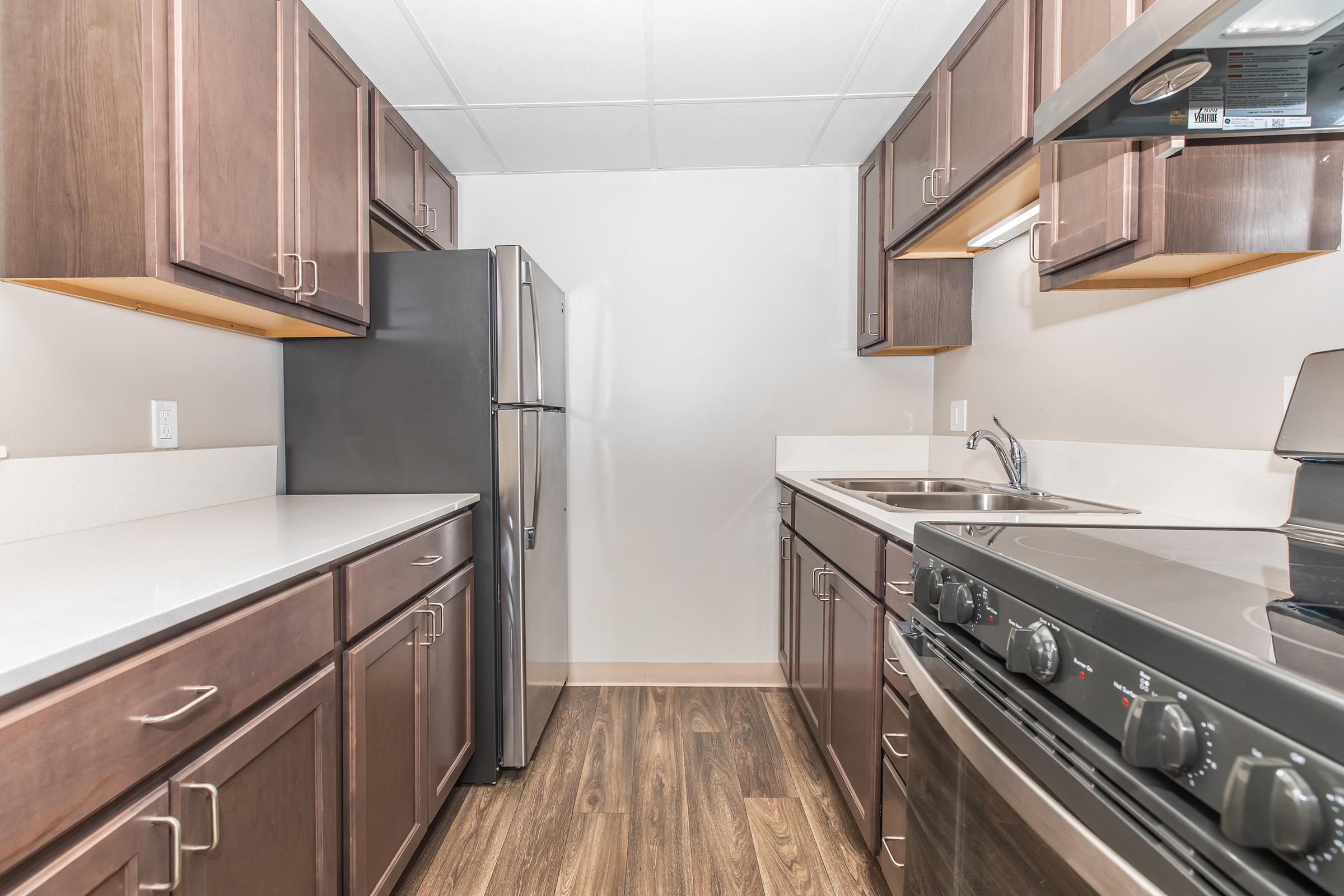Modern kitchen featuring dark wood cabinetry, stainless steel appliances including a refrigerator and oven, a sink, and light countertops. The flooring is a warm wood-like laminate, and the walls are painted in a neutral tone. Natural light brightens the space, enhancing the contemporary design.