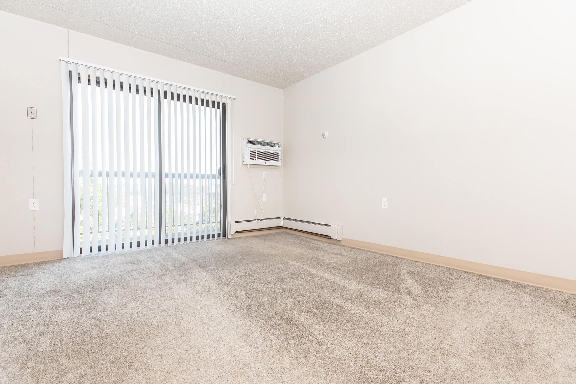 A spacious, empty room with light-colored walls and beige carpeting. There are vertical blinds covering a sliding glass door that leads to a balcony, and an air conditioning unit mounted on the wall. The room is well-lit and has a clean, minimalistic appearance.
