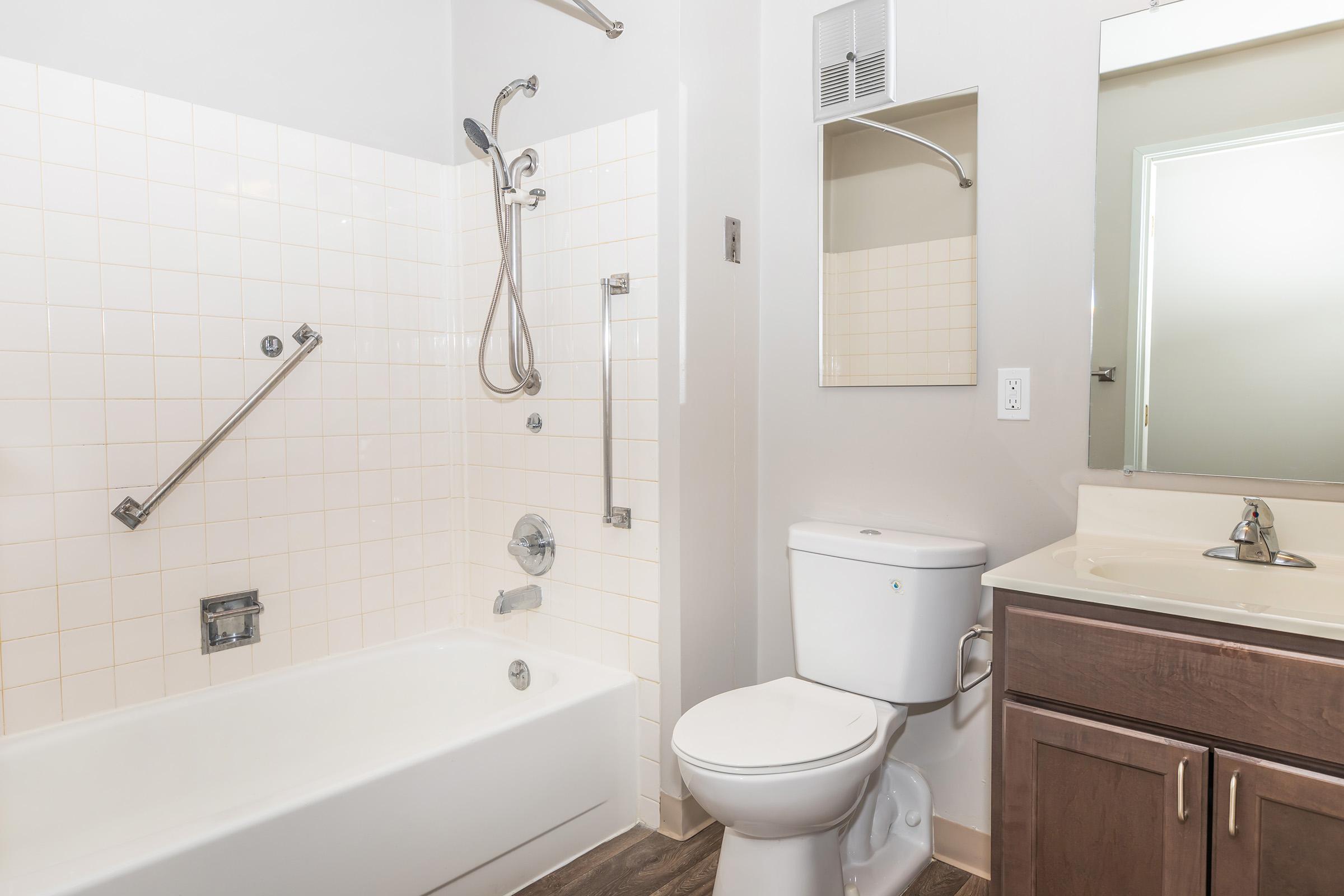 A clean and modern bathroom featuring a white bathtub with a shower attachment, a toilet, a single sink with a mirror above it, and light-colored tiled walls. The floor has a wood-like finish, and the space is well-lit with natural light coming from a nearby window.