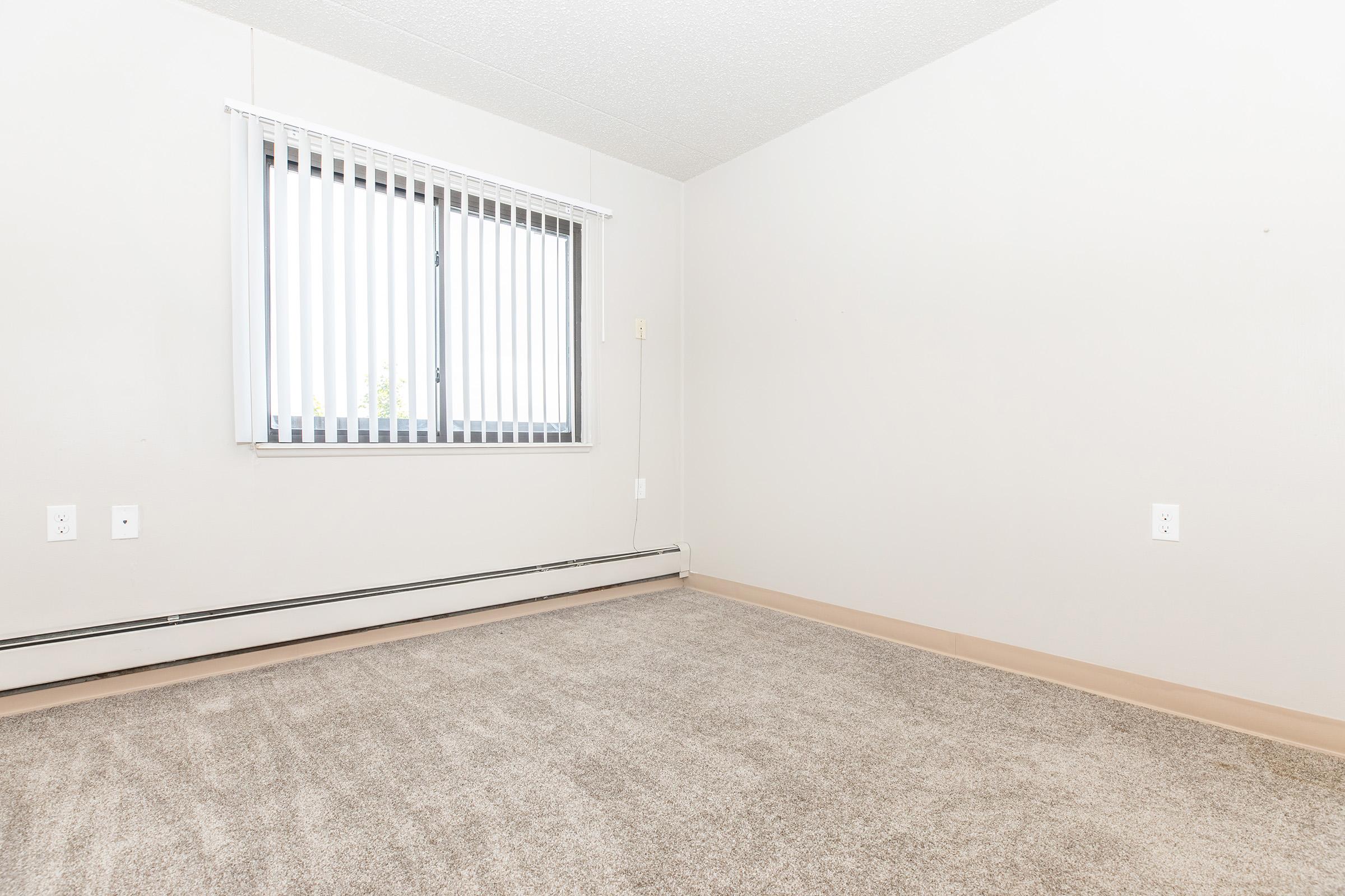 A vacant room with light beige walls and carpet. There is a window with vertical blinds on the left side, allowing natural light to enter. The room features baseboard heating along the wall. No furniture or decorations are present, creating a minimalistic and empty appearance.