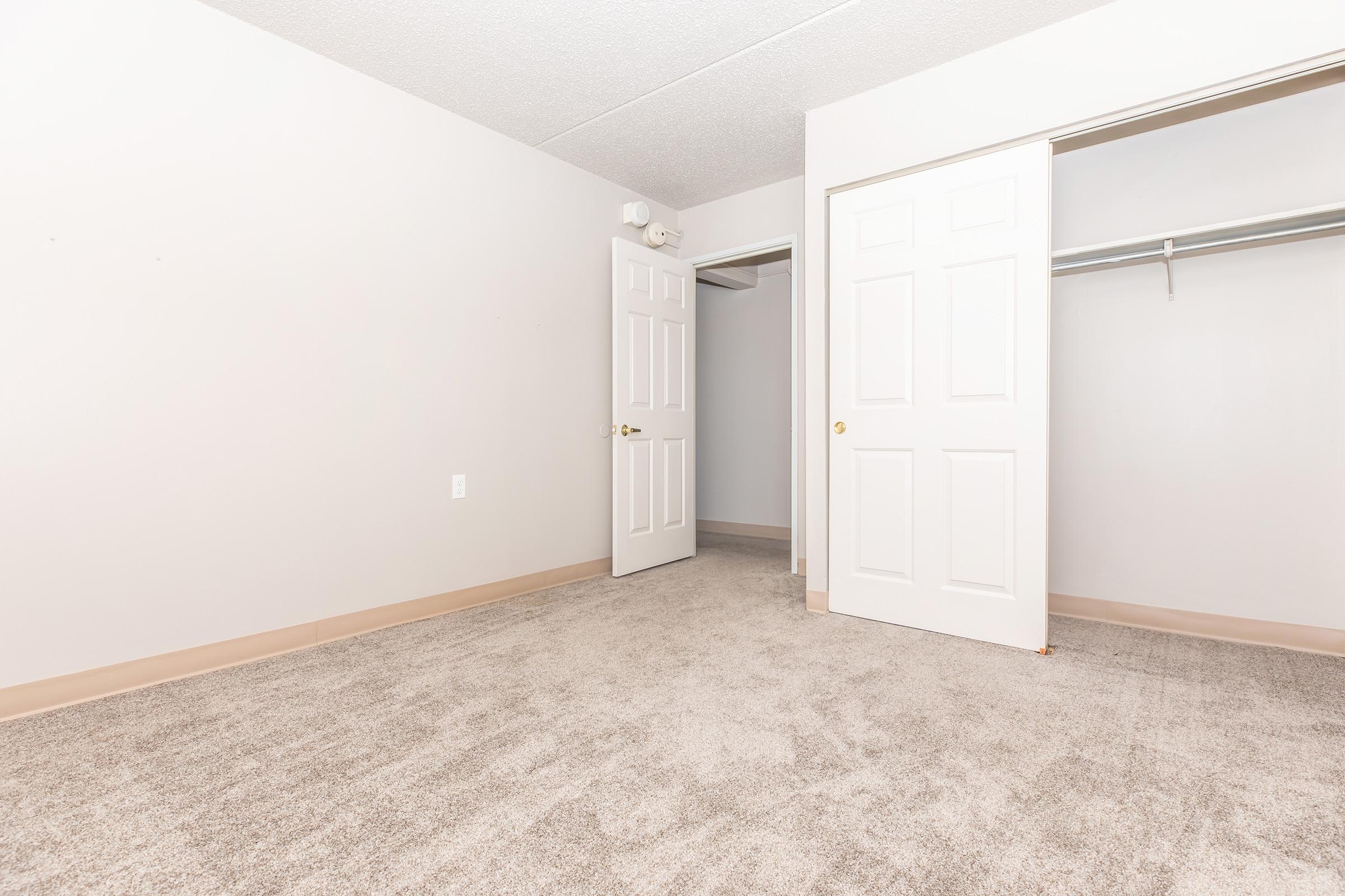 An empty room with light gray walls and beige carpet. Two open doors lead to a closet, which has a hanging rod. The space is well-lit and unfurnished, giving a clean and spacious appearance.