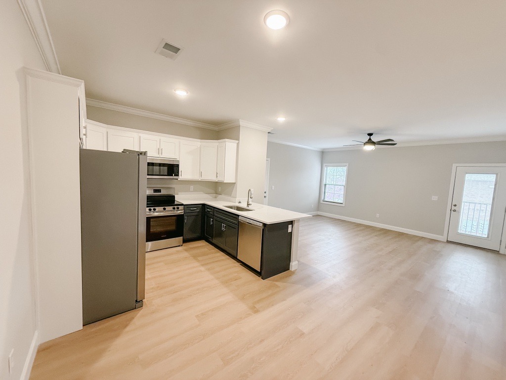 Modern kitchen and living area featuring stainless steel appliances, white cabinets, and a large window. The space has light wooden flooring and a ceiling fan, with an adjacent door leading outside. Bright, minimalistic design with ample natural light.