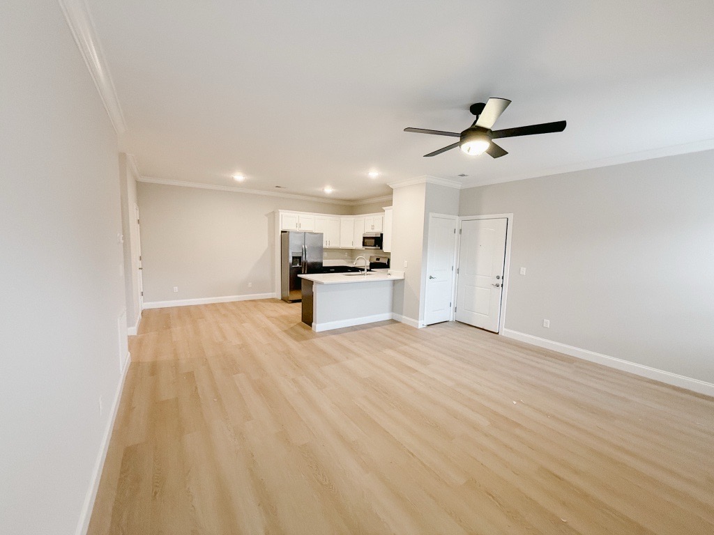 A spacious, empty living area featuring light-colored walls and wooden flooring. A ceiling fan is visible, and the kitchen can be seen in the background, equipped with white cabinets and modern appliances. The area appears well-lit and ready for furnishings.