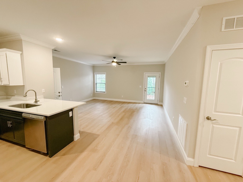 A spacious, light-filled living area with pale hardwood floors. The kitchen features white cabinets and a stainless steel sink. A ceiling fan hangs over the main area, and a door on the right leads to an outdoor space. Walls are painted neutral tones, enhancing the airy feel of the room.