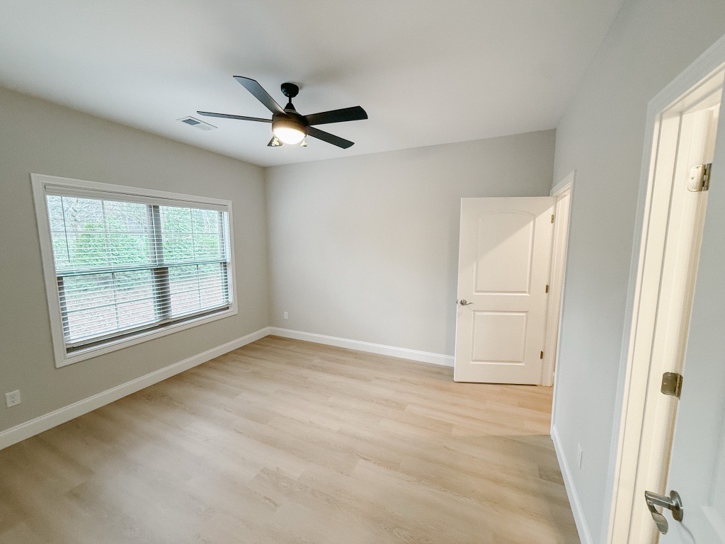 A well-lit bedroom featuring light-colored walls and wooden flooring. The space includes a ceiling fan, a large window with horizontal blinds, and an open door leading to another room. The overall ambiance is modern and minimalistic, with natural light illuminating the room.