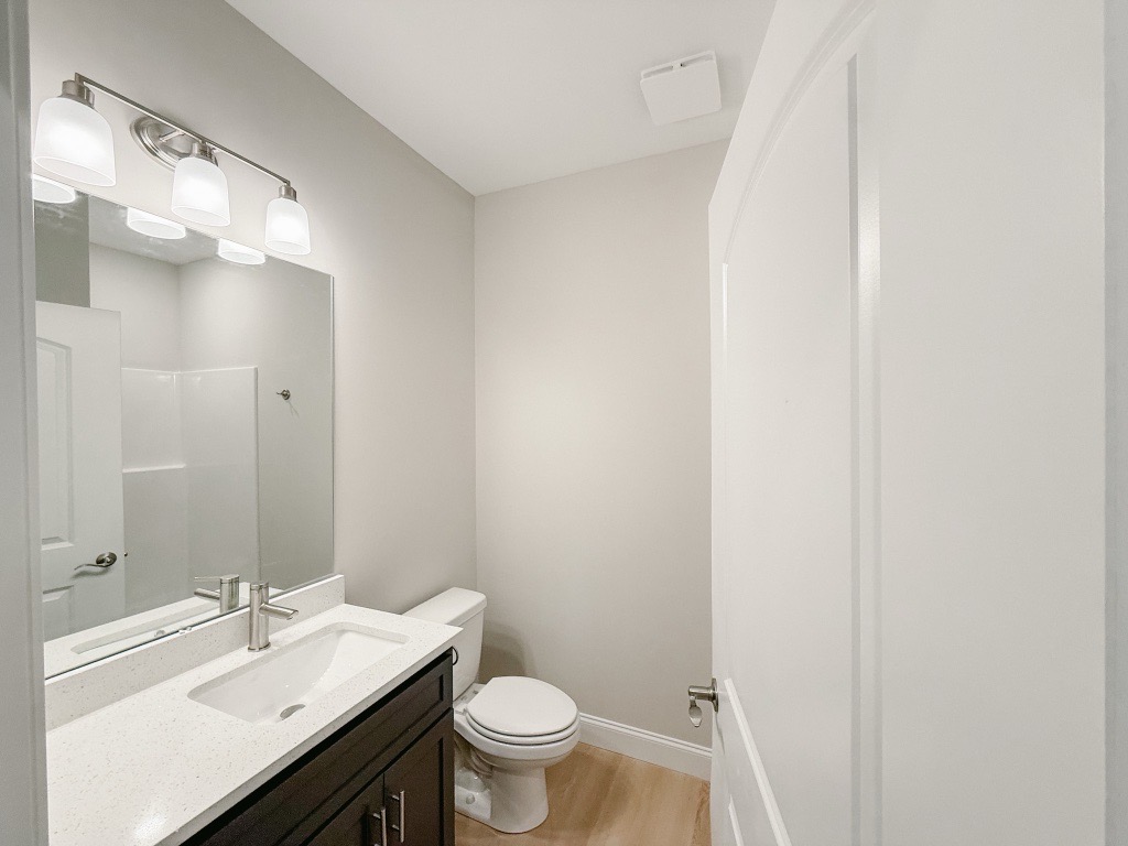 A modern bathroom featuring a white countertop with a sink, a mirror above it, and a toilet. The walls are painted a light gray, and there is a shower stall visible in the background. The flooring is a light wood. The lighting consists of three pendant-style fixtures above the sink.