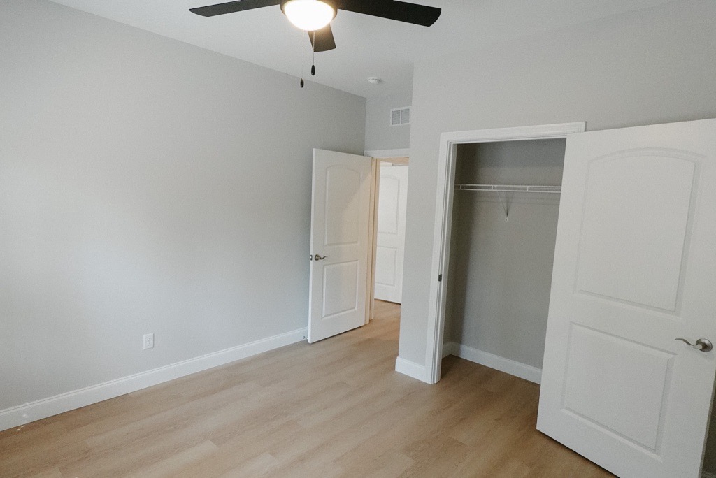 A clean, empty room with light gray walls and a ceiling fan. There are two open doors leading to closets, and the flooring is light wood. The room is well-lit, giving a spacious and modern feel, suitable for potential decoration or use as a bedroom or office.