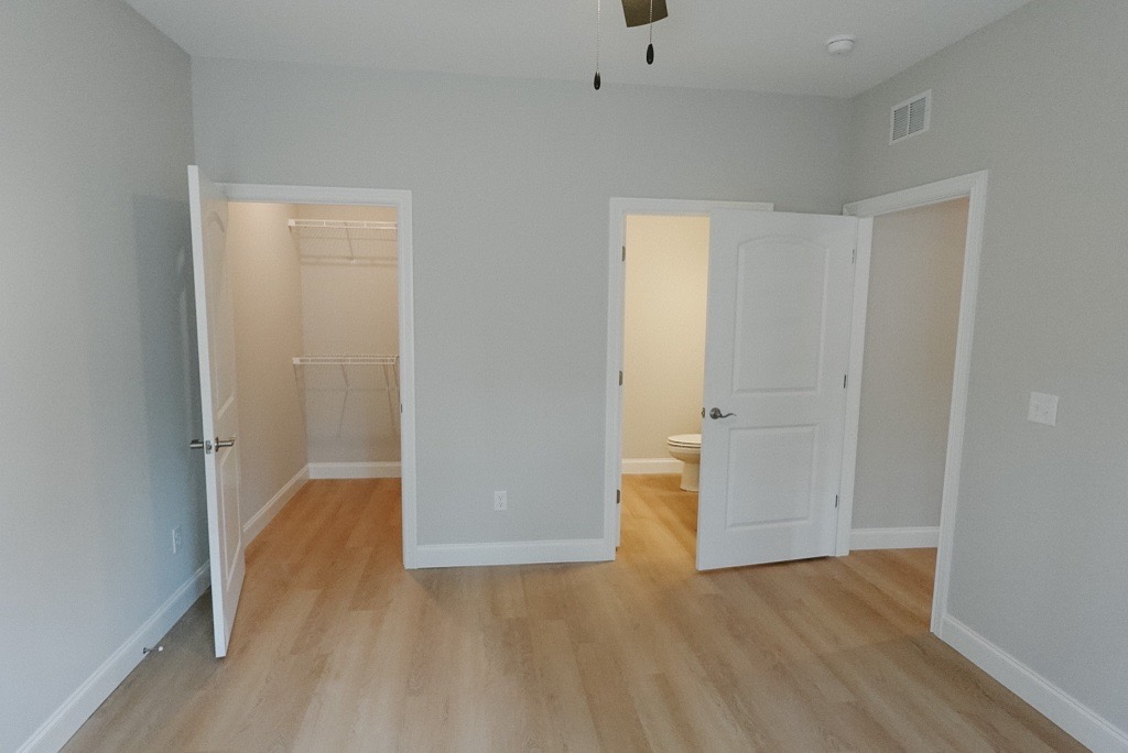 A light-colored room featuring beige walls and wood laminate flooring. The image shows two doorways: one leading to a spacious closet with a hanging rack and the other to a bathroom. A ceiling fan hangs in the center of the room, providing an airy feel. The room is well-lit with natural light.