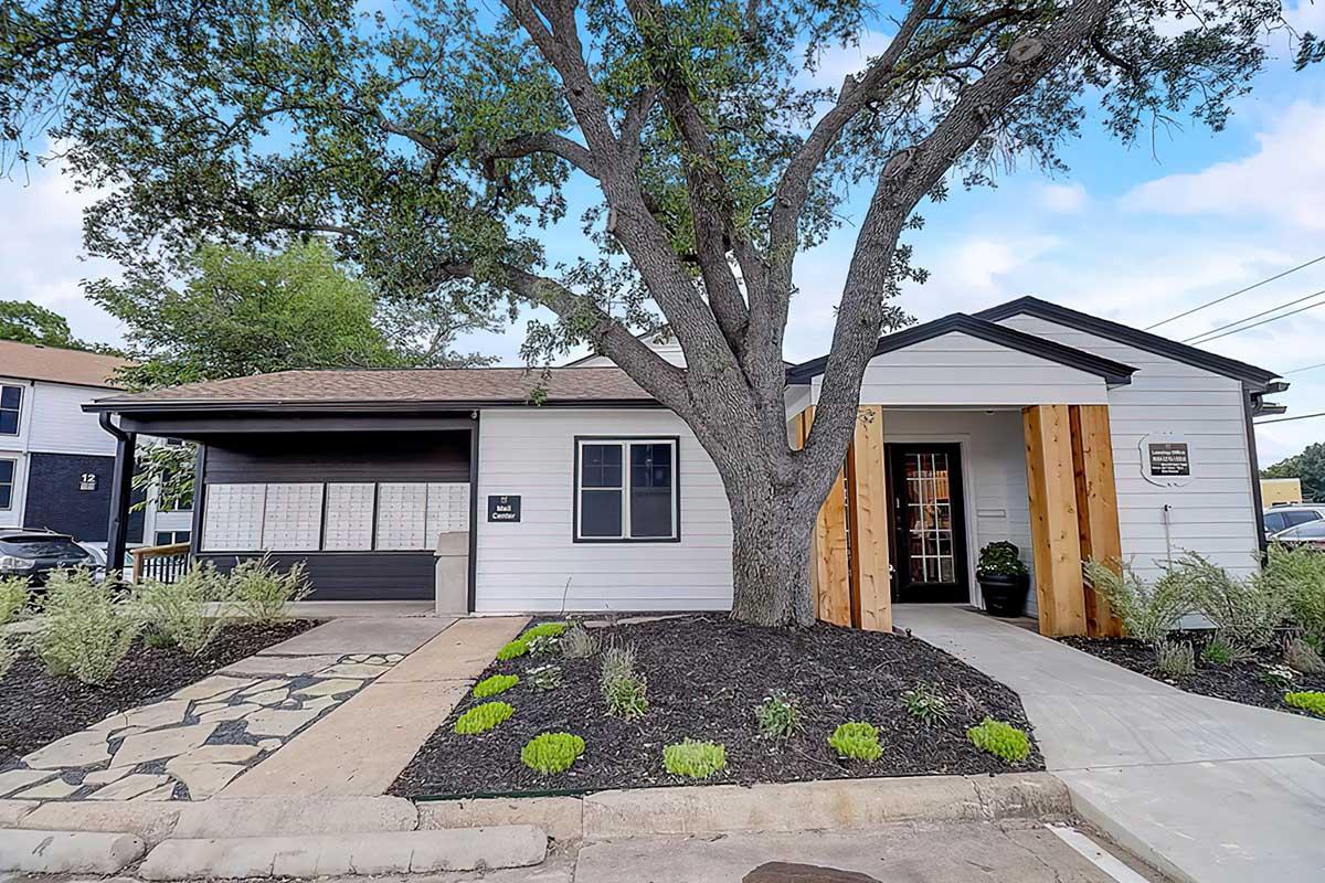 A modern single-story building with white siding, wooden accents, and a spacious entrance. The front yard features decorative landscaping with plants, a tree, and a stone pathway leading to the entrance. The property is well-maintained and inviting, set against a clear blue sky.