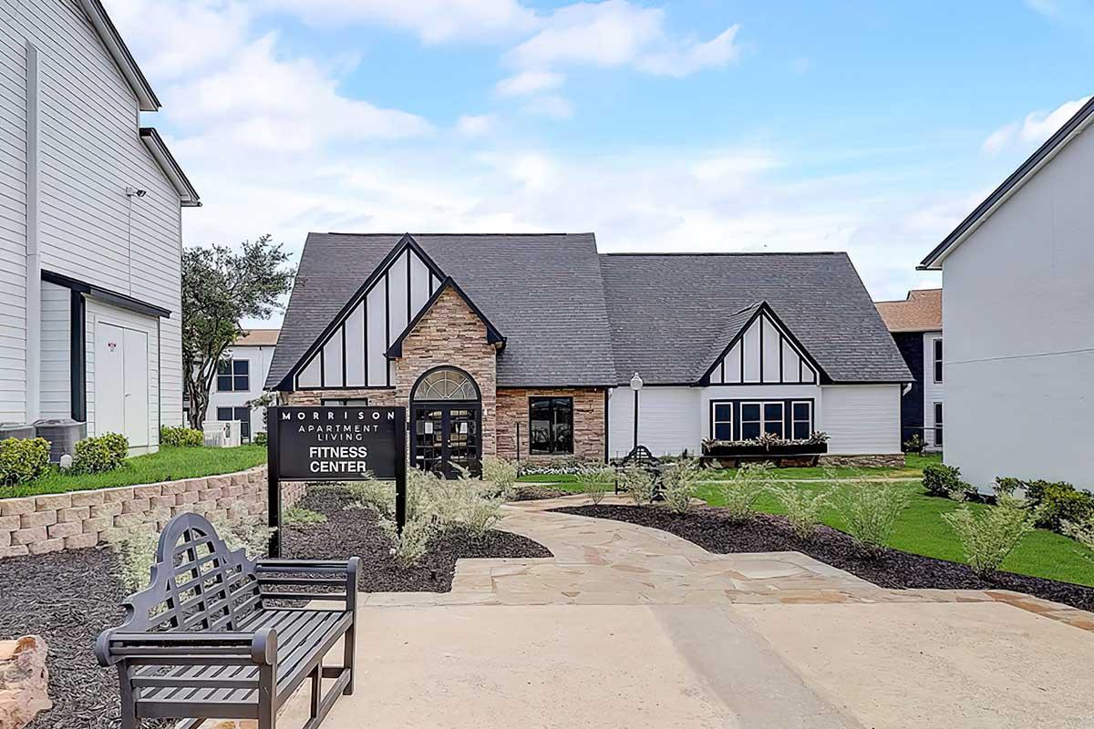 Exterior view of the Morrison Apartment Fitness Center, featuring a gray and tan building with a pitched roof, large windows, and a stone facade. A sign indicating the fitness center is at the forefront, with landscaped areas including shrubs and a path leading to the entrance. A bench sits nearby.