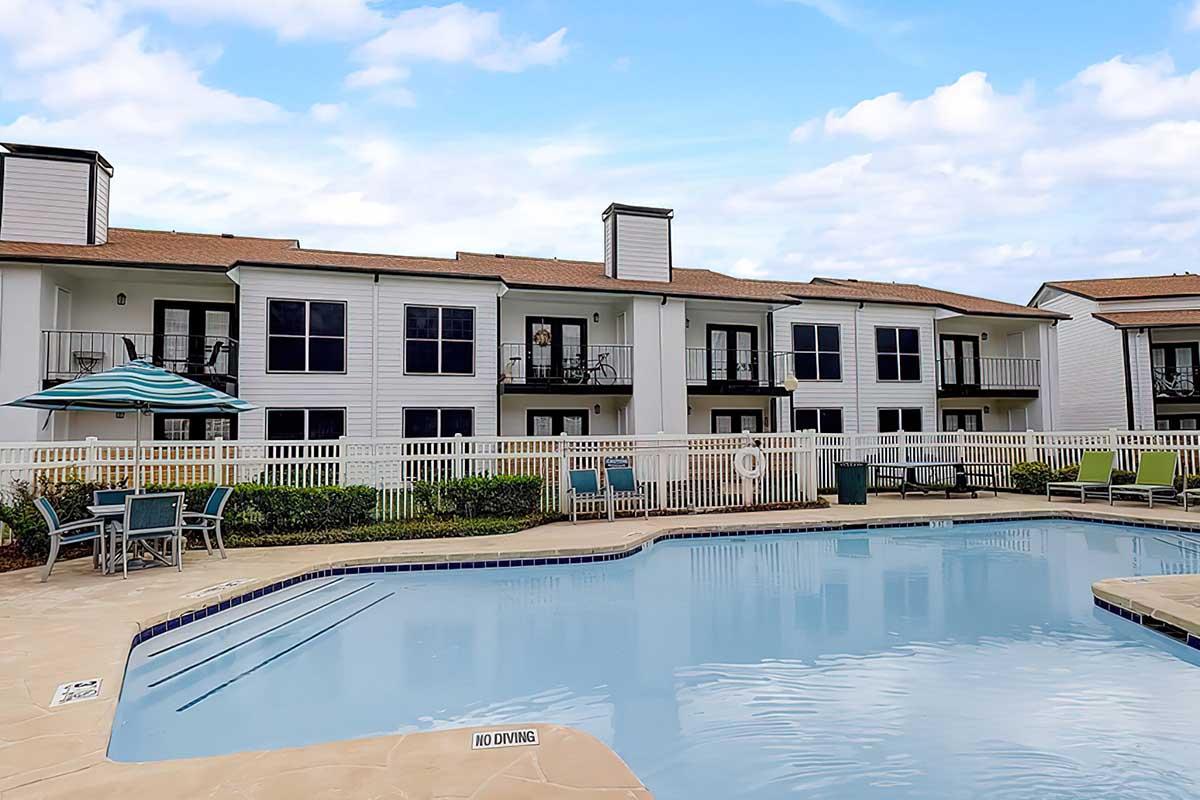 A tranquil outdoor scene featuring a swimming pool surrounded by lounge chairs and tables. In the background, there are well-maintained apartment buildings with balconies. A large umbrella provides shade, and a āNo Divingā sign is visible near the poolās edge. The sky is partly cloudy, suggesting a pleasant day.