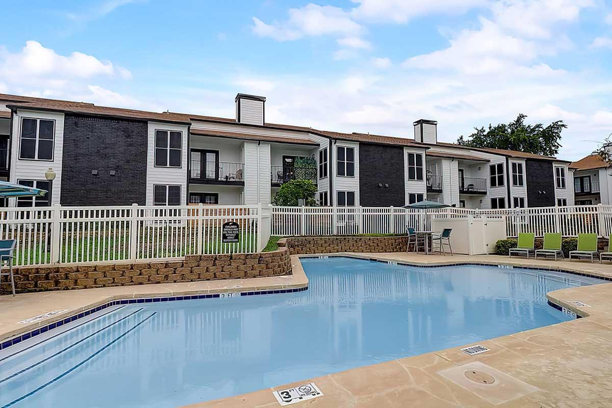 A swimming pool surrounded by lounge chairs and a landscaped area, with several apartment buildings in the background. The sky is partly cloudy, creating a bright and inviting outdoor space.