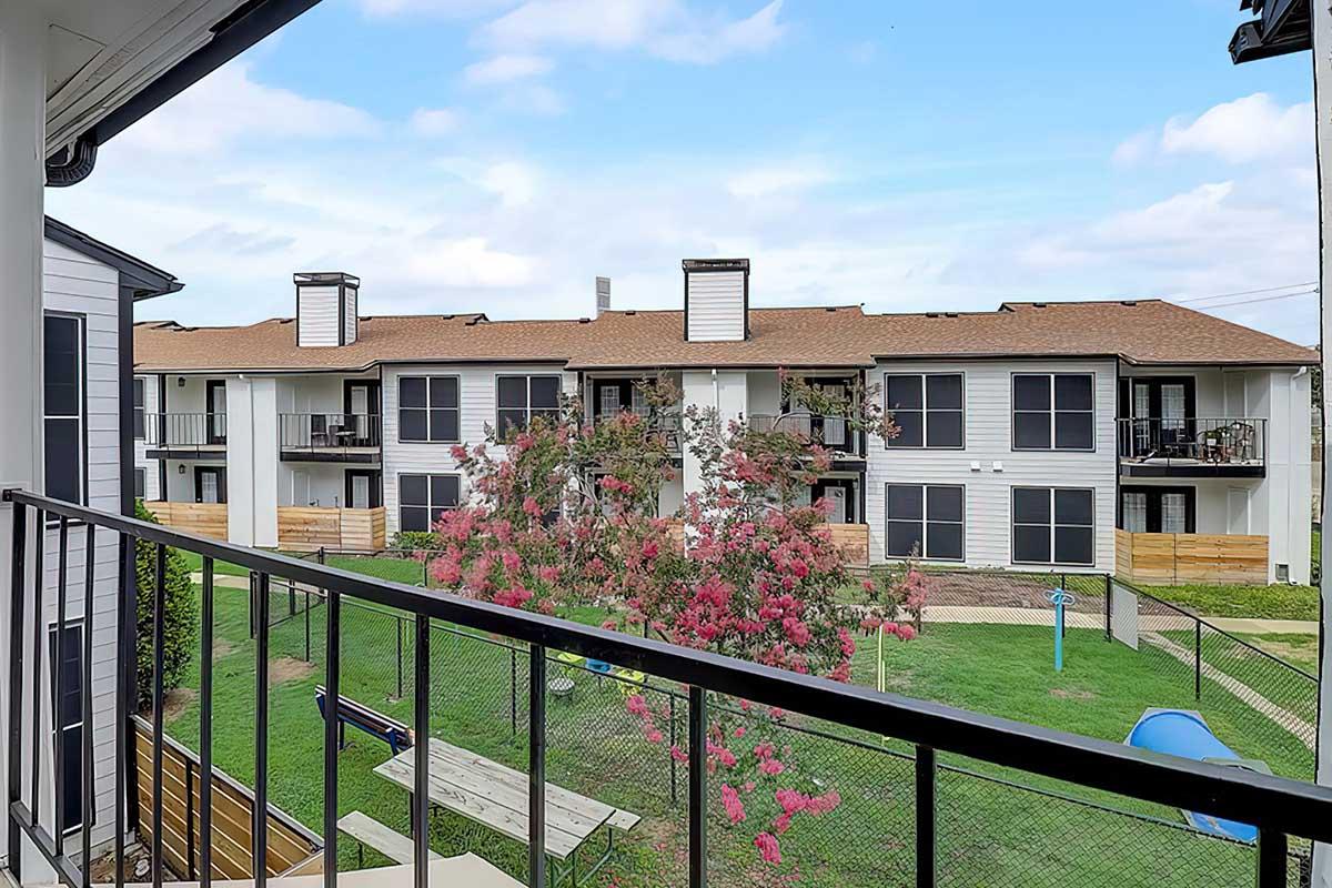 View from a balcony overlooking an apartment complex. The scene features a grassy area, a flowering tree, and several buildings with balconies. The sky is partly cloudy, and there are fences and outdoor seating visible in the surroundings.