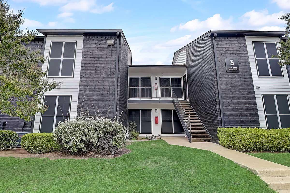 A modern two-story apartment building with a dark exterior. The building features balconies, multiple windows, and a central entrance with stairs leading to the second floor. Surrounding the building is a well-maintained green lawn and shrubs. Clear blue sky in the background.