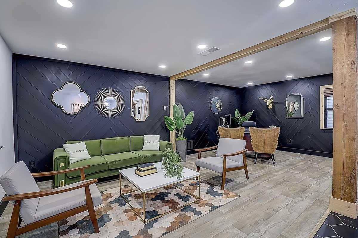 Modern living room featuring a dark accented wall with decorative mirrors, a green sofa, and two simple armchairs. Thereās a light-colored coffee table on a patterned rug, with potted plants adding a touch of greenery. Wood beams and flooring give a warm, inviting feel to the space.