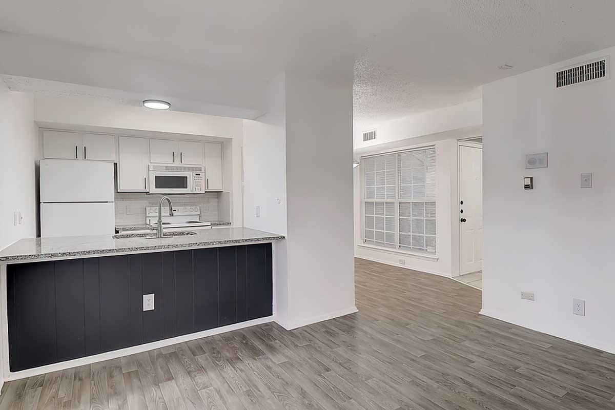 Modern kitchen with white cabinets, a microwave, and a refrigerator, featuring a granite countertop that overlooks a bright living space. The adjacent area includes large windows and a door leading outside, showcasing a clean and minimalist design with light-colored walls and wood-style flooring.