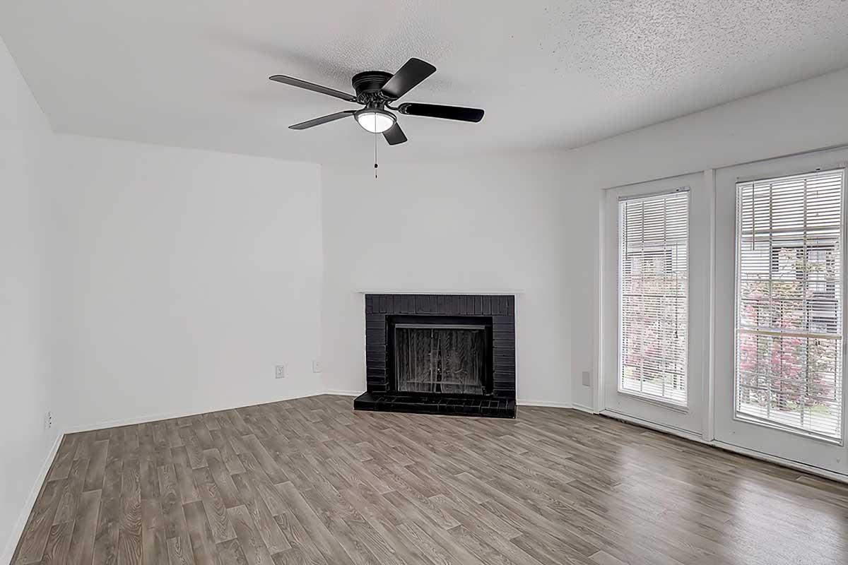 Empty living room with light-colored walls and wood-style flooring. Features a ceiling fan, a black fireplace surrounded by a black mantel, and two large windows with blinds. Natural light is entering the room.