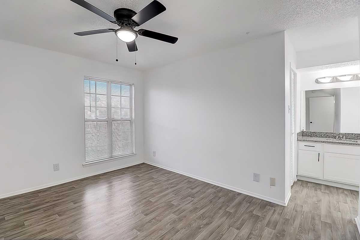 A bright, minimally furnished room with a wooden floor, featuring a ceiling fan and a large window bringing in natural light. On one side, there is a small bathroom area with a countertop and mirror visible, creating an open and airy atmosphere.