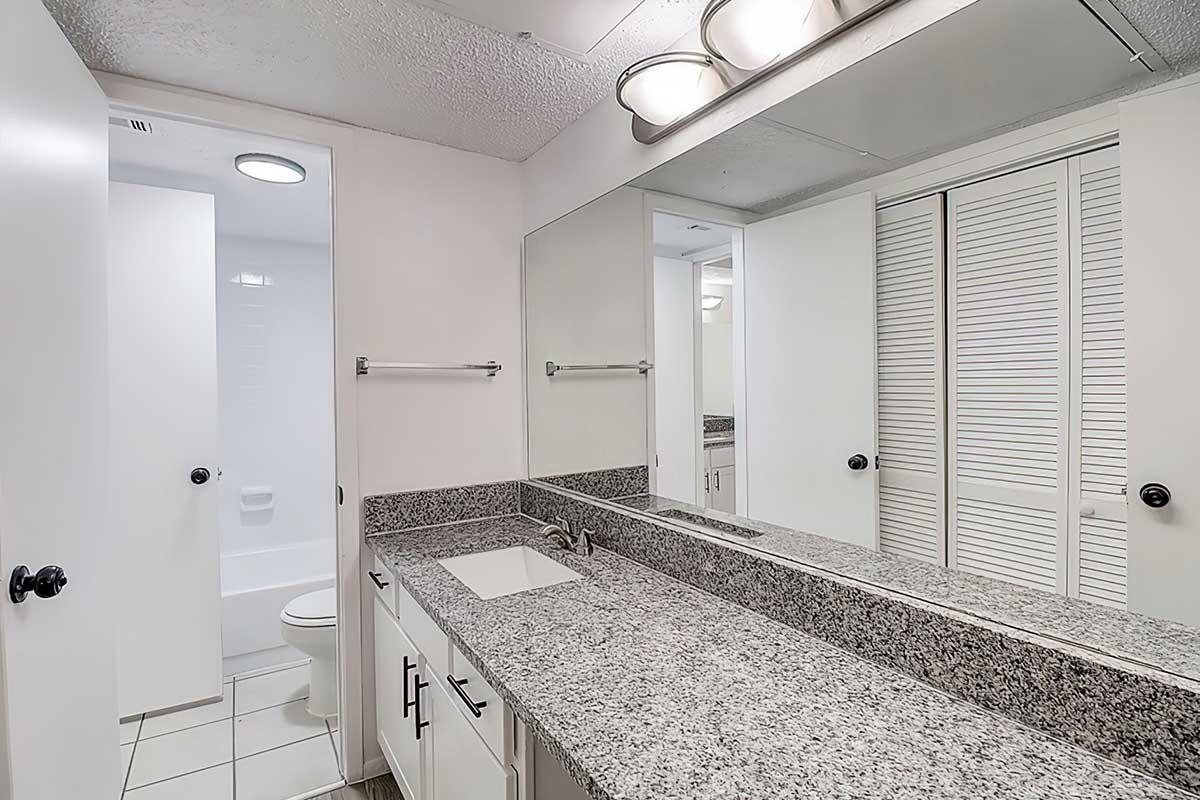 A modern bathroom featuring a granite countertop with an integrated sink, large mirror, and stylish lighting. White cabinets and a closet with sliding doors are visible. A toilet is partially seen through an open door in the background, along with white tiled flooring and bright lighting.