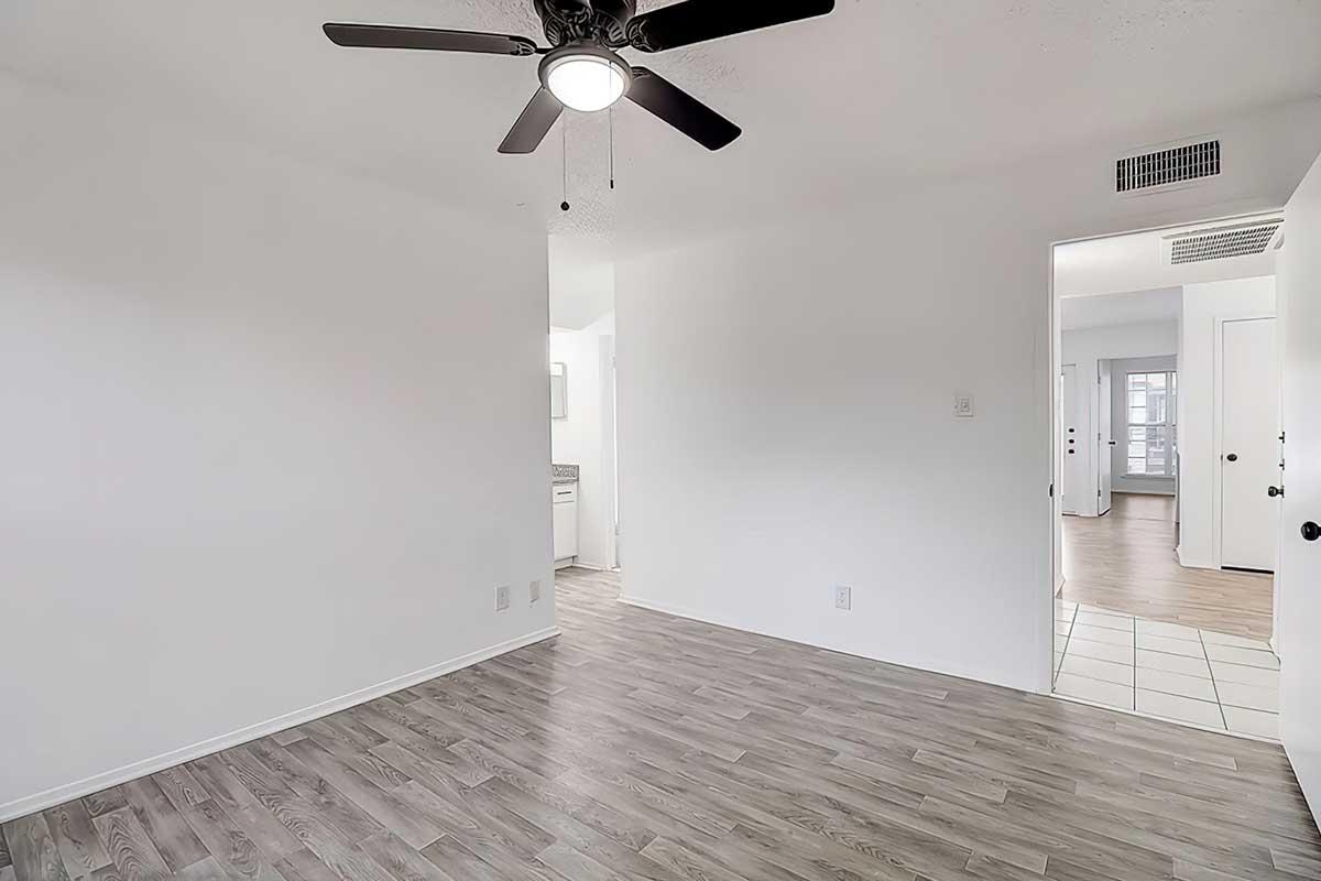 Interior view of a bright room featuring light-colored walls and a ceiling fan. The floor is covered with light wood laminate. A doorway visible in the background leads to a bathroom, while another doorway opens to a hallway with additional rooms.