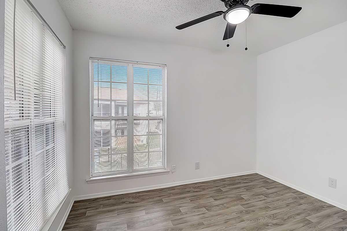 A bright, empty room with light-colored walls and a ceiling fan. There are large windows with blinds allowing natural light to enter, and the floor is covered with wood-like laminate. The space appears ready for occupancy or decoration.