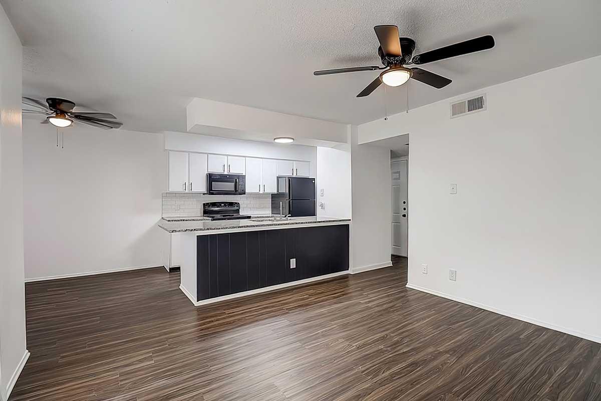 A modern open-concept kitchen and living area featuring dark wood flooring, black appliances, and a breakfast bar. There are two ceiling fans, and the space is brightly lit with white walls, creating a clean and inviting atmosphere. The layout promotes easy movement between the kitchen and living space.