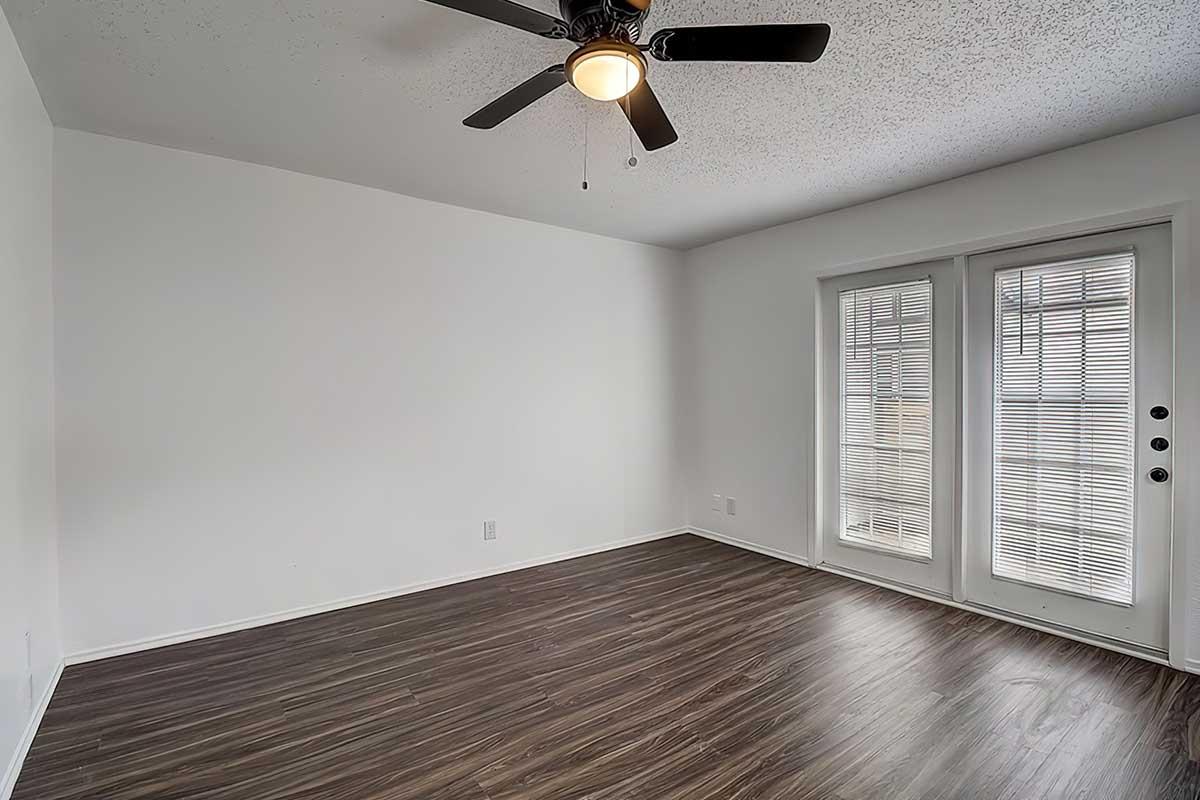 Empty room with light-colored walls and dark wooden flooring. A ceiling fan with three blades is mounted on the ceiling. There are sliding glass doors with vertical blinds on one side, providing natural light. The space appears clean and ready for furnishing.