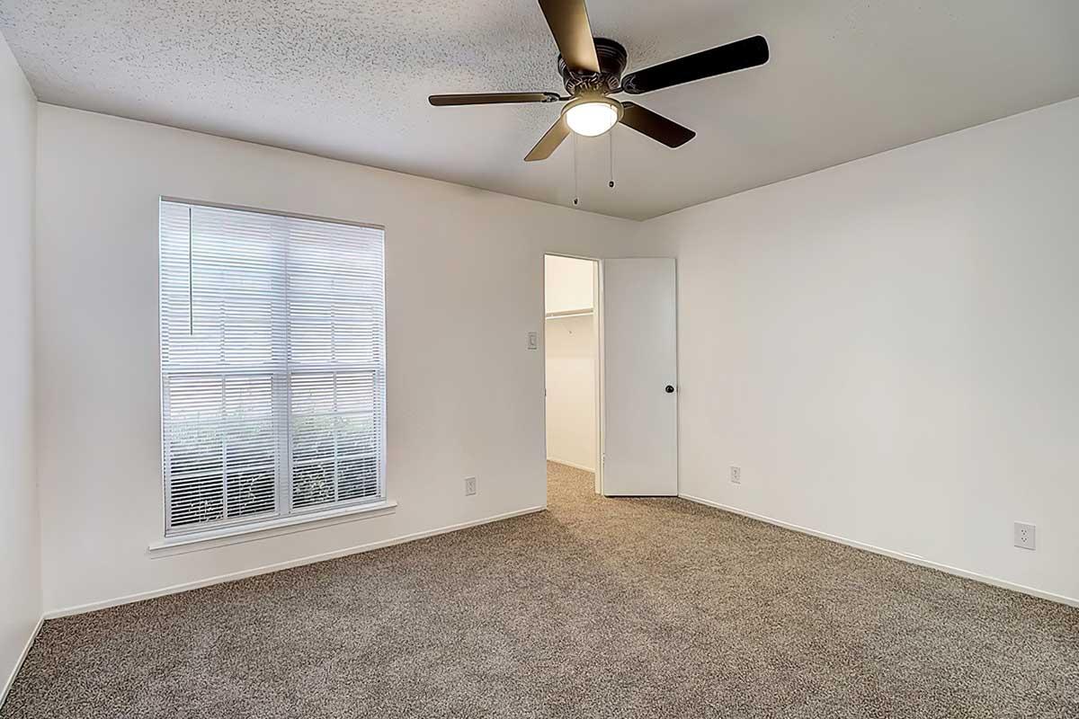 A bright, empty room with light-colored walls and beige carpet. It features a ceiling fan with five blades, a window with white blinds, and an open door leading to a closet. The room is spacious and well-lit, creating an inviting atmosphere.